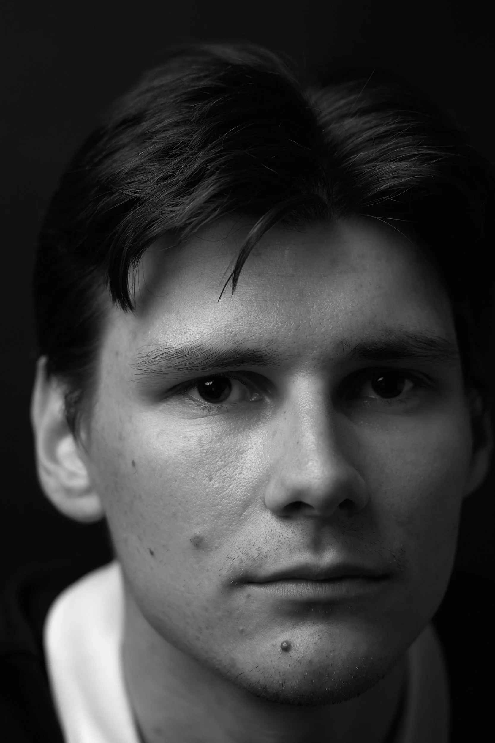Schwarz-weiß-Fotografie eines Jungen mit dunklen Haaren, sichtbarer Hauttextur, manchmal unscharf um den Mund, mit einem dunklen Hintergrund.