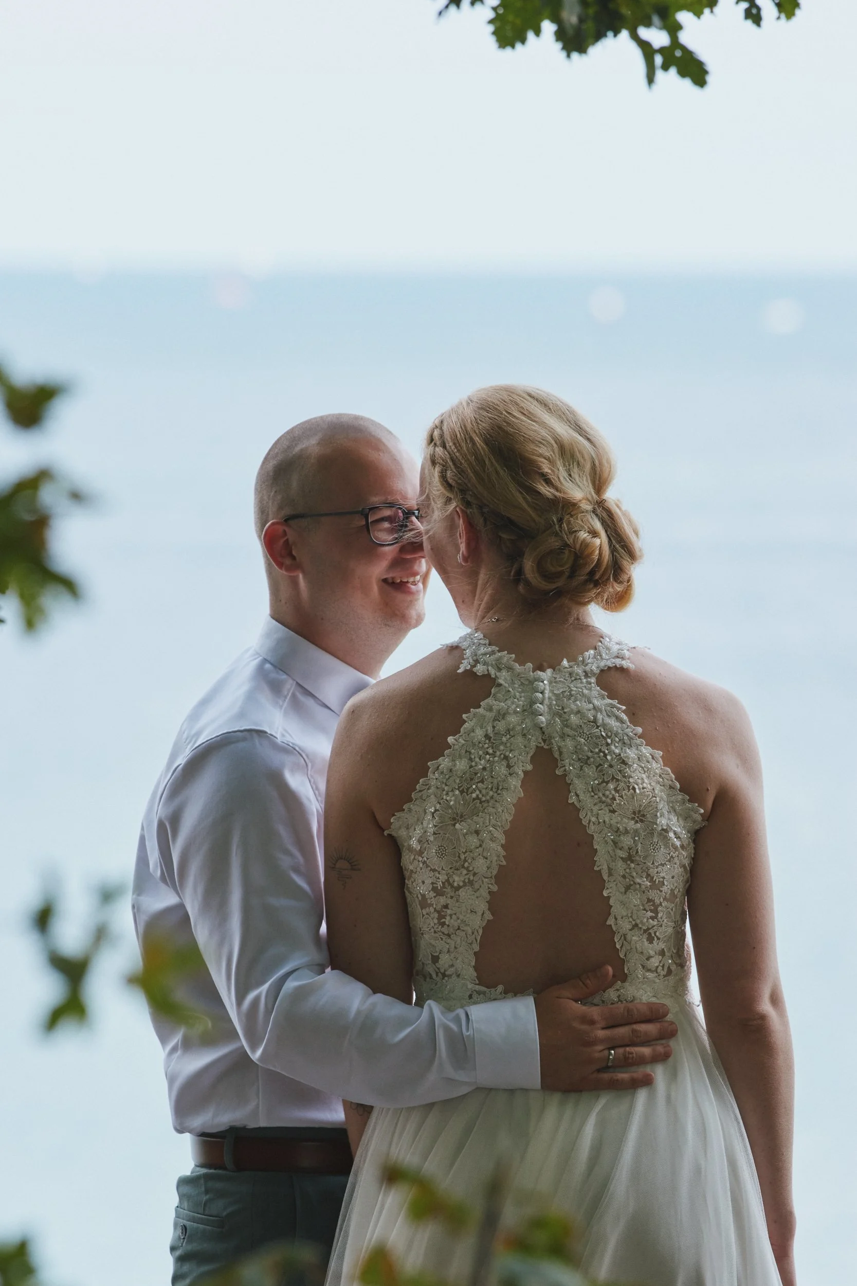 Ein Brautpaar steht sich bei einem Hochzeitsfoto im Freien gegenüber, umarmt sich und lächelt einander an, im Hintergrund sieht man das Meer und den Himmel.