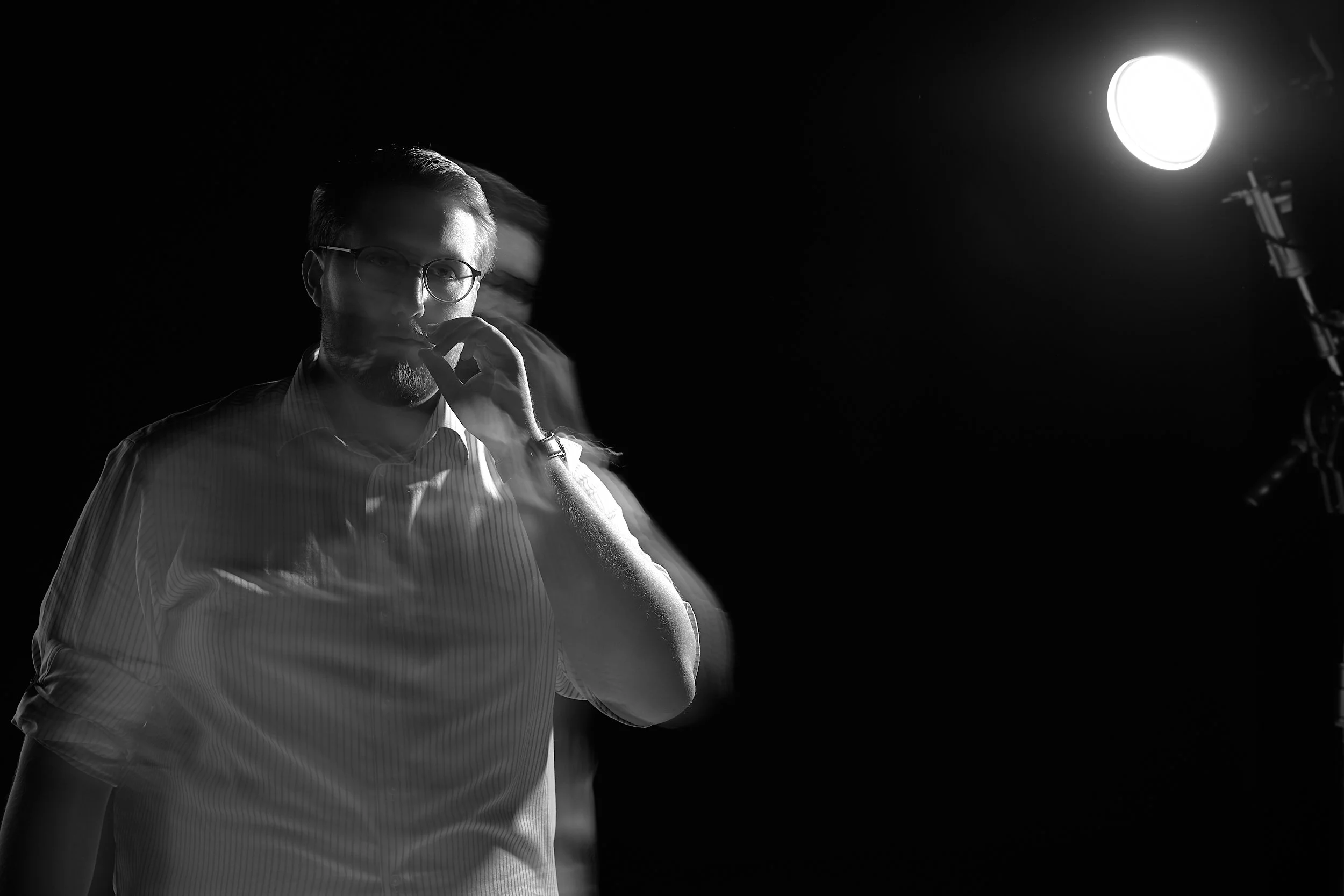 Schwarz-weiß-Fotografie eines Mannes mit Brille, der nachdenklich aussieht, während er eine Hand vor den Mund hält, im Hintergrund ein helles Licht.