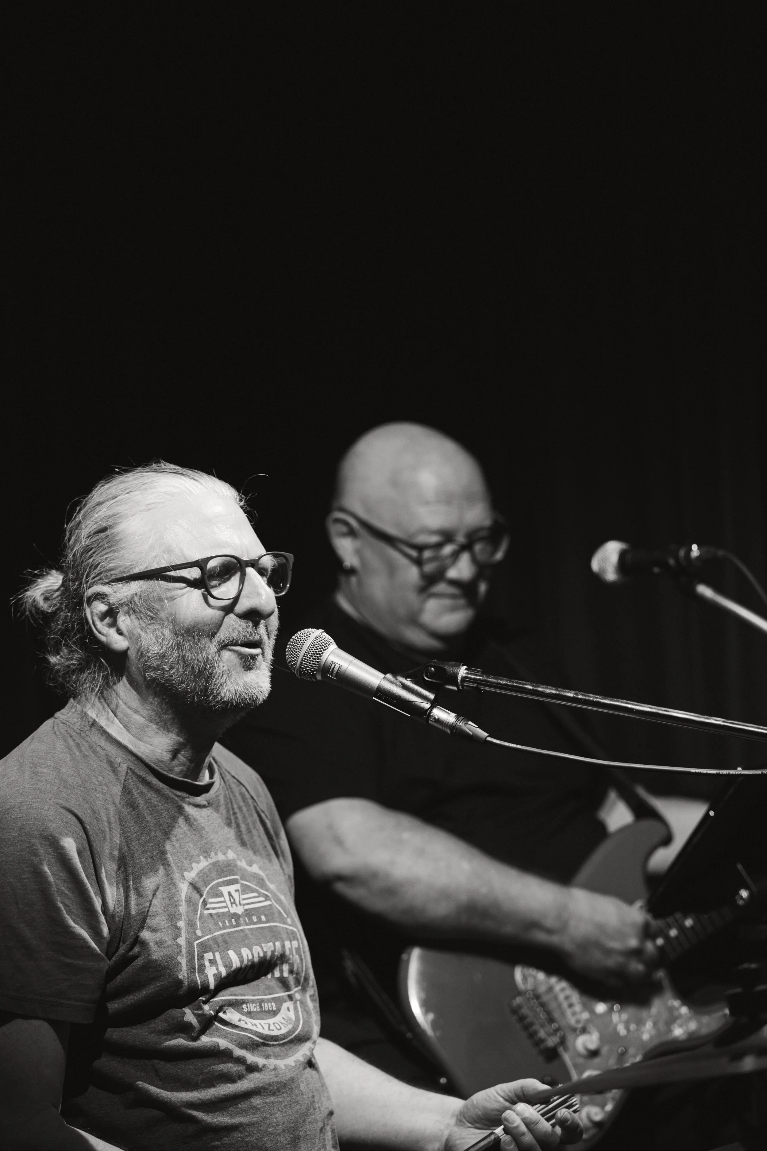 Zwei Männer beim Musizieren: Der ältere Mann mit Brille und grauem Haar spielt Gitarre, der andere Mann mit Glatze und ebenfalls Brille singt in ein Mikrofon, beide sitzen an einem Konzert. Es ist eine schwarze und weiße Fotografie.