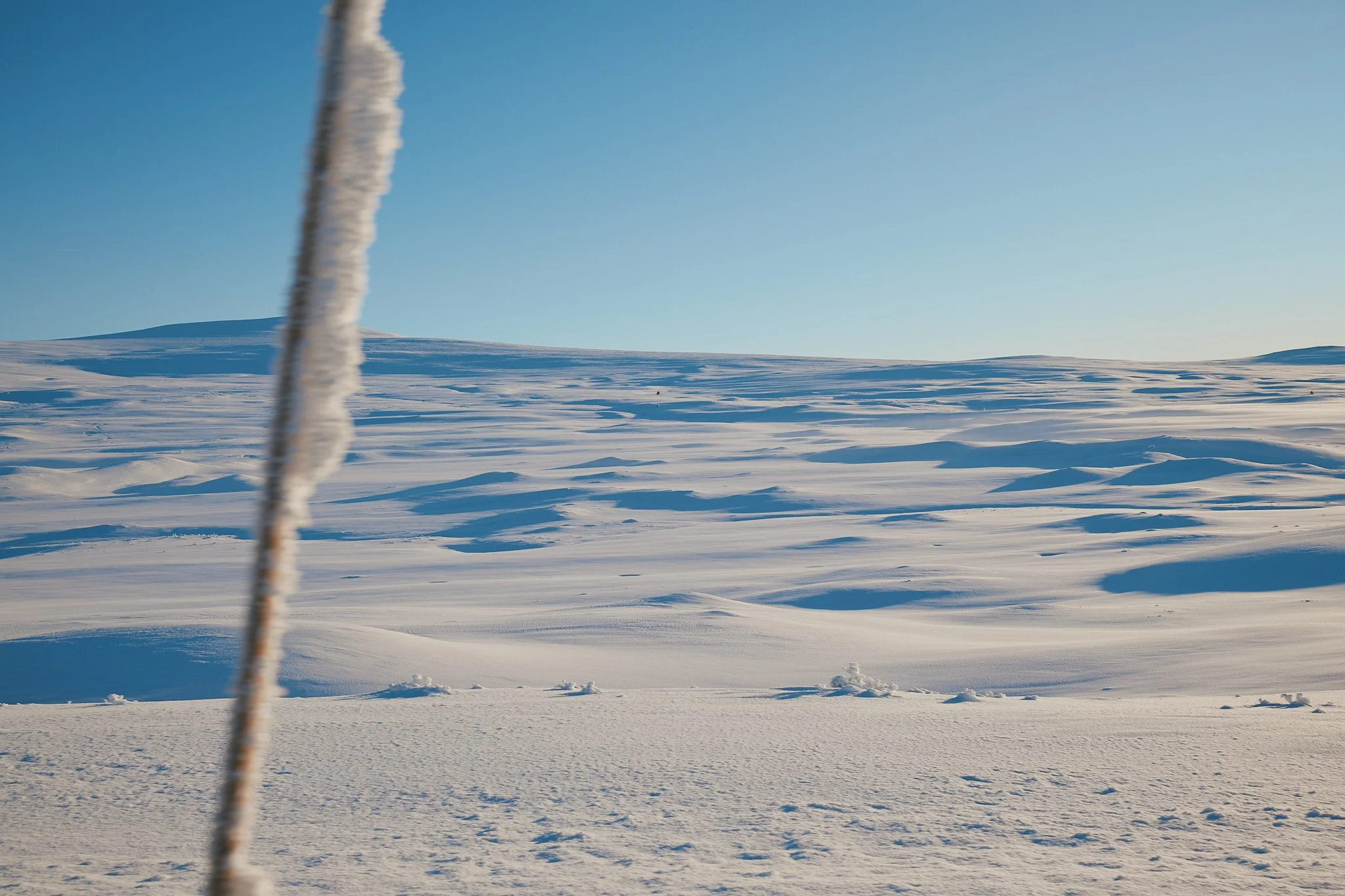 Schneebedecktes, offenes Landschaft mit Hügeln unter klarem blauen Himmel, im Vordergrund ein verschneiter Pfad und ein vertikaler, verunklappter Gegenstand.