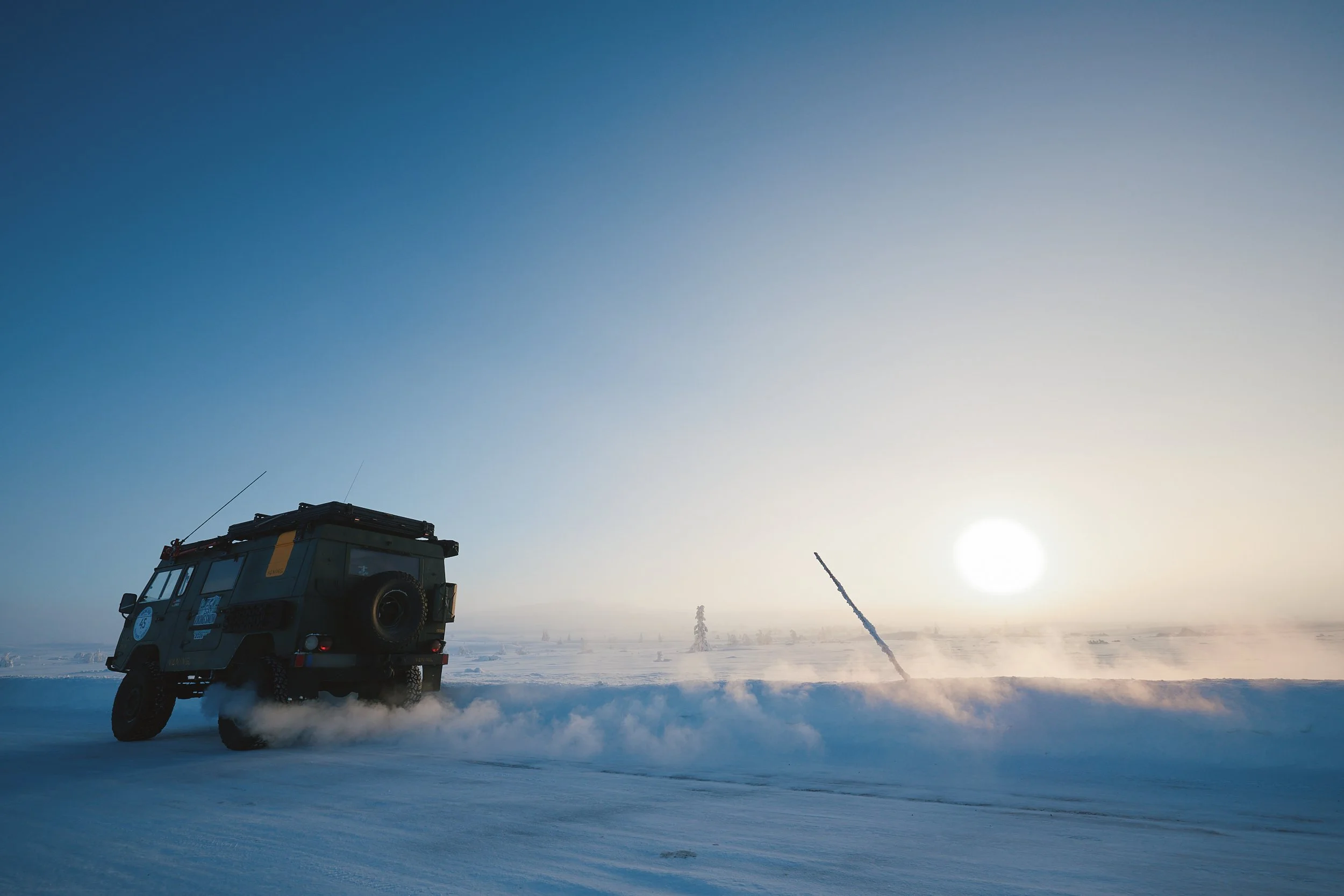 Ein schwarzer Geländewagen fährt durch verschneiten Boden, während die Sonne am Horizont untergeht, und der Himmel ist klar mit wenigen Wolken.