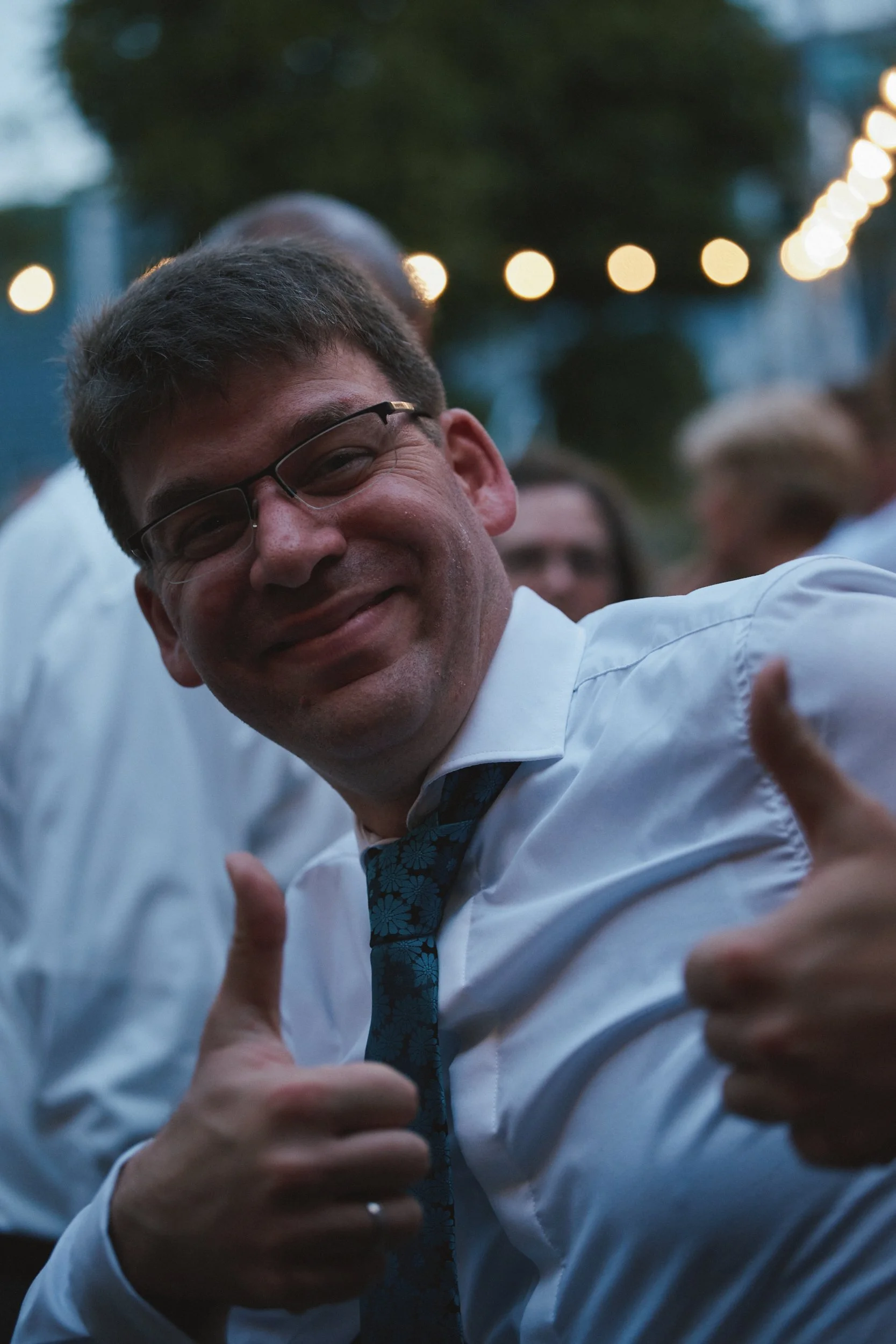 Mann mit Brille und Krawatte, der lächelt und Daumen zeigt, bei einer festlichen Veranstaltung im Freien, im Hintergrund Lichterketten.