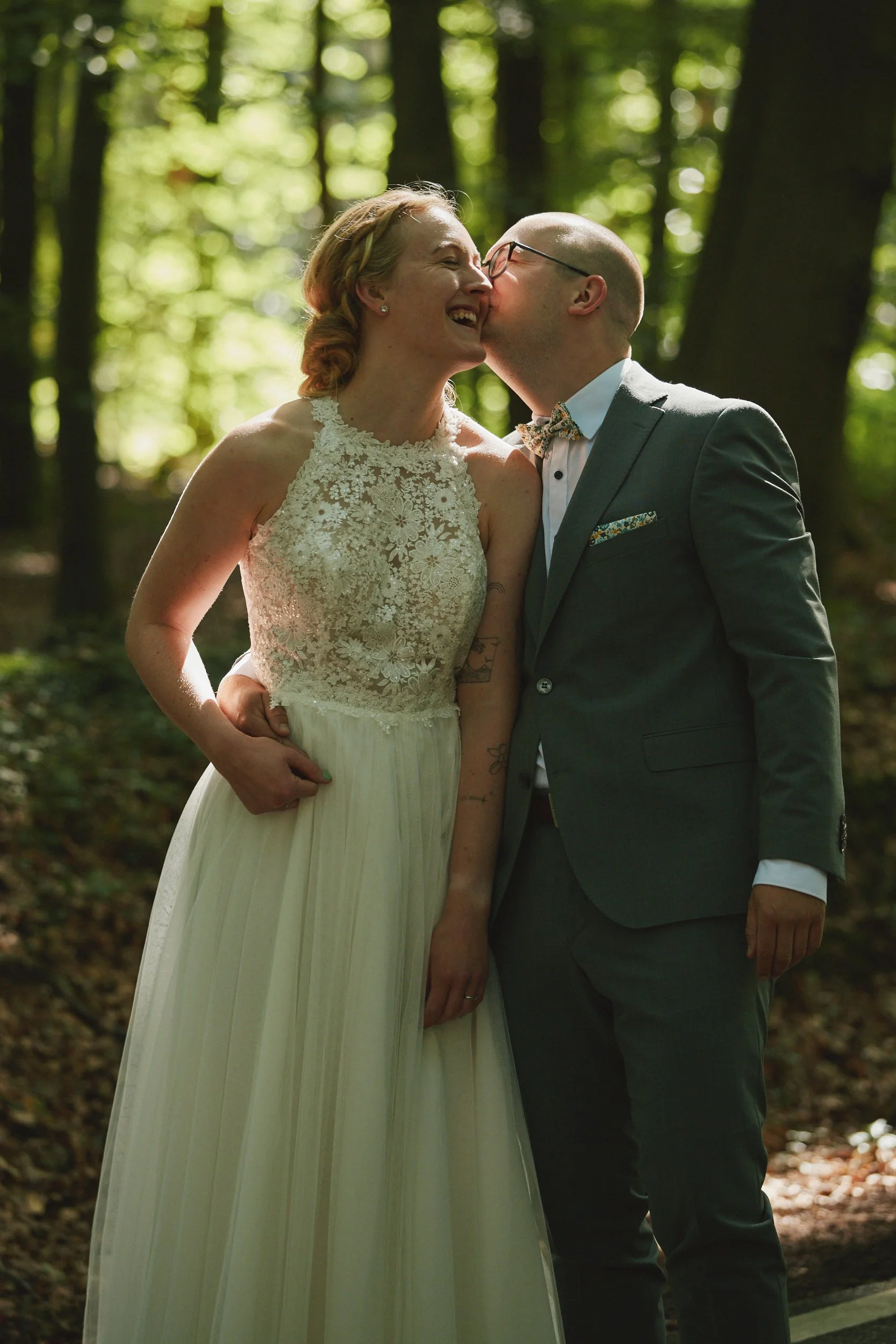 Ein lachendes Paar bei einer Hochzeit im Wald, der Mann küsst die Frau auf die Wange.
