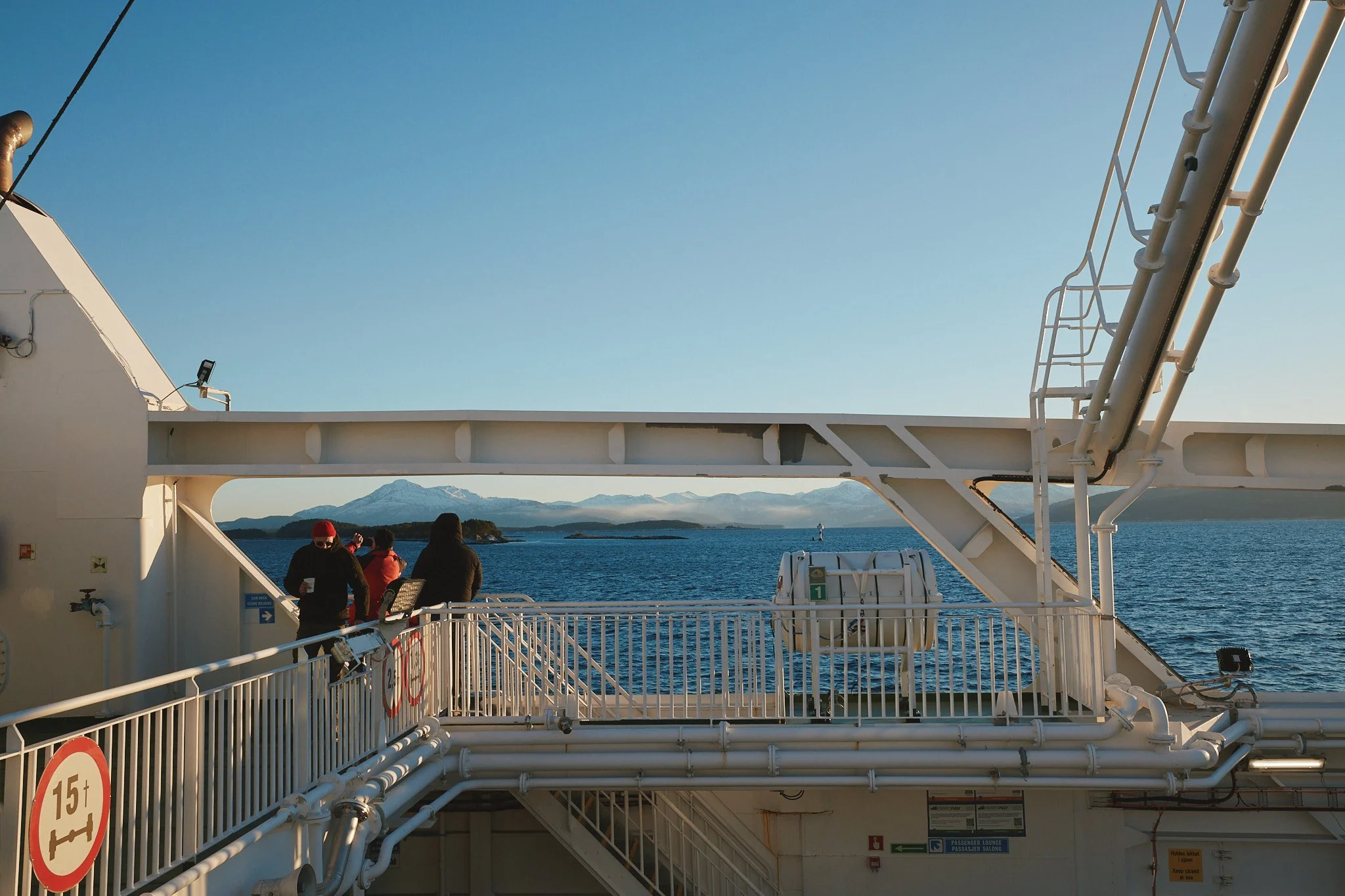 Menschen stehen auf der oberen Deckebene eines Kreuzfahrtschiffes vor einem Meer mit Bergen im Hintergrund, während die Sonne scheint.