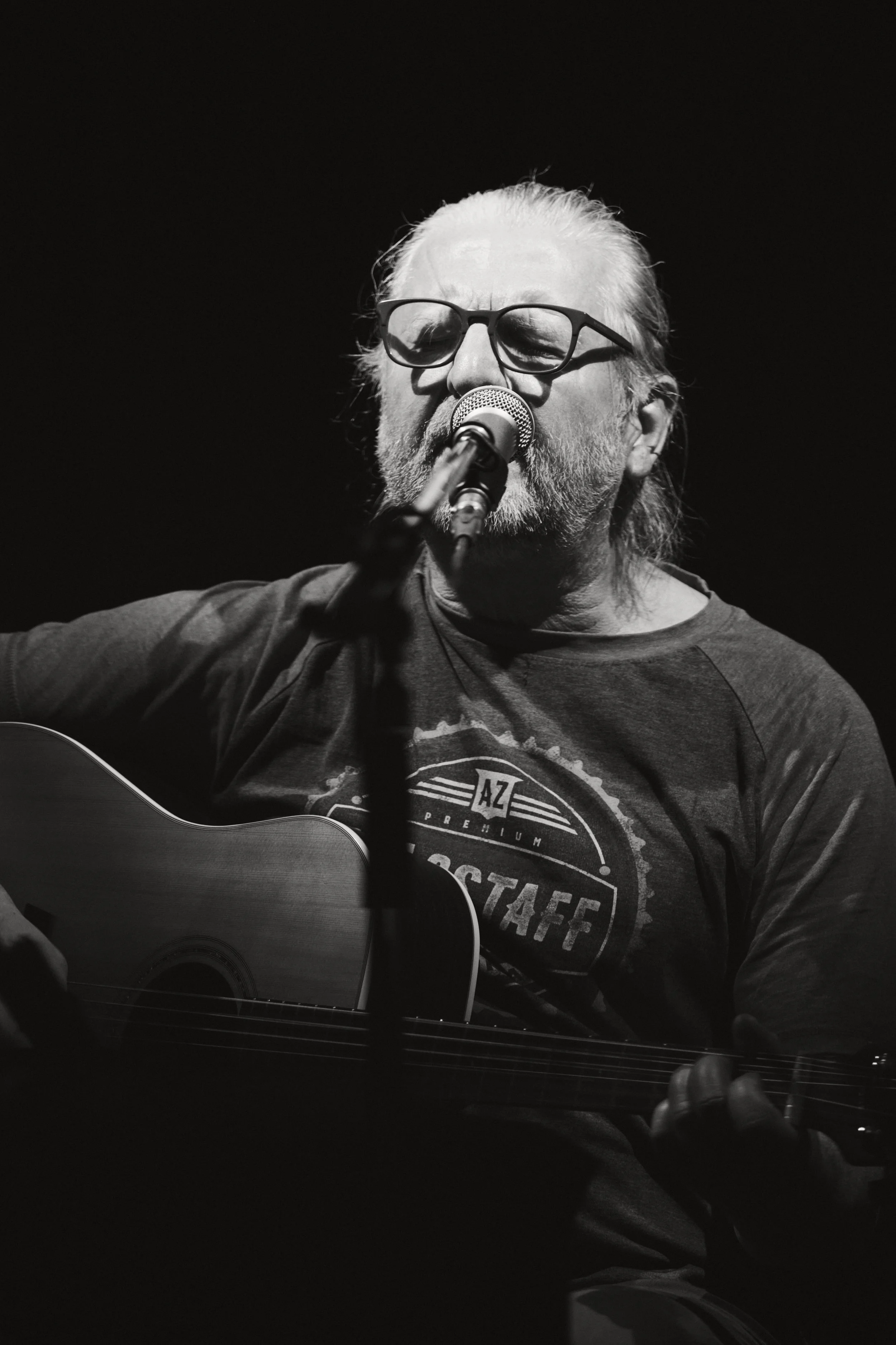 Schwarz-weiß-Foto eines älteren Mannes mit Brille, der singt und auf einer Gitarre spielt.