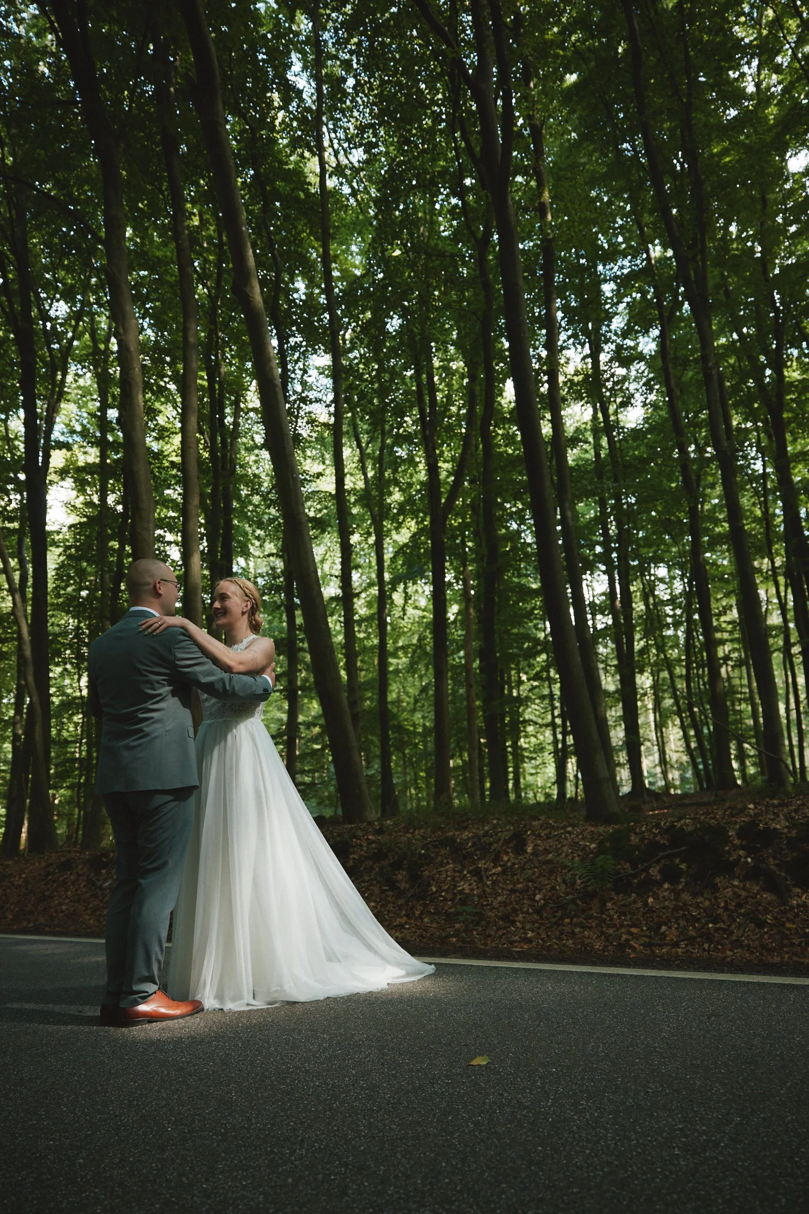 Ein Brautpaar tanzt auf einer Straße, umgeben von einem grünen Wald. Der Mann trägt einen grauen Anzug, die Frau ein weißes Hochzeitskleid.