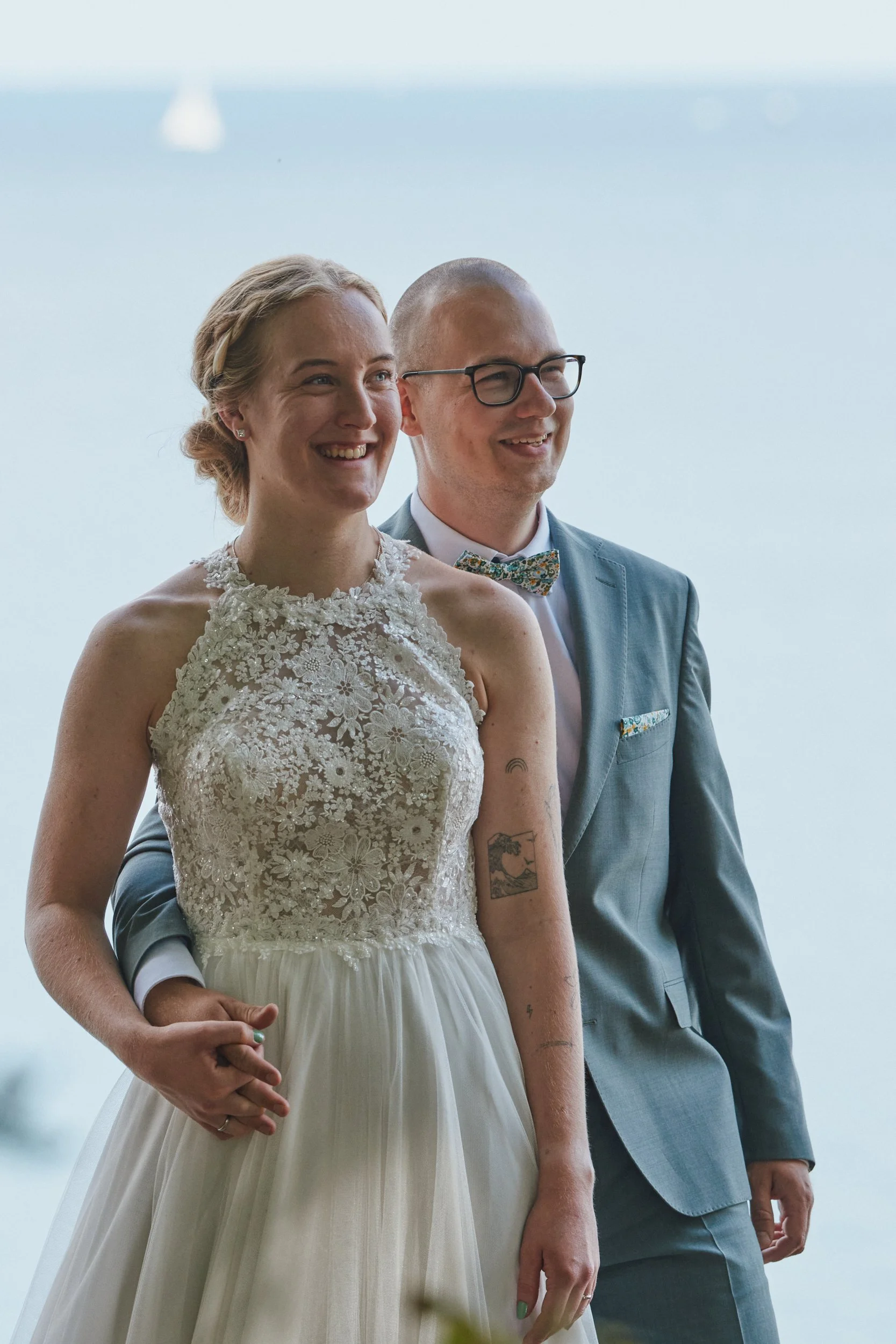 Ein Brautpaar bei einer Hochzeit, die Braut trägt ein weißes Kleid mit Blumenmuster und der Bräutigam trägt einen grauen Anzug mit Fliege und Brille.