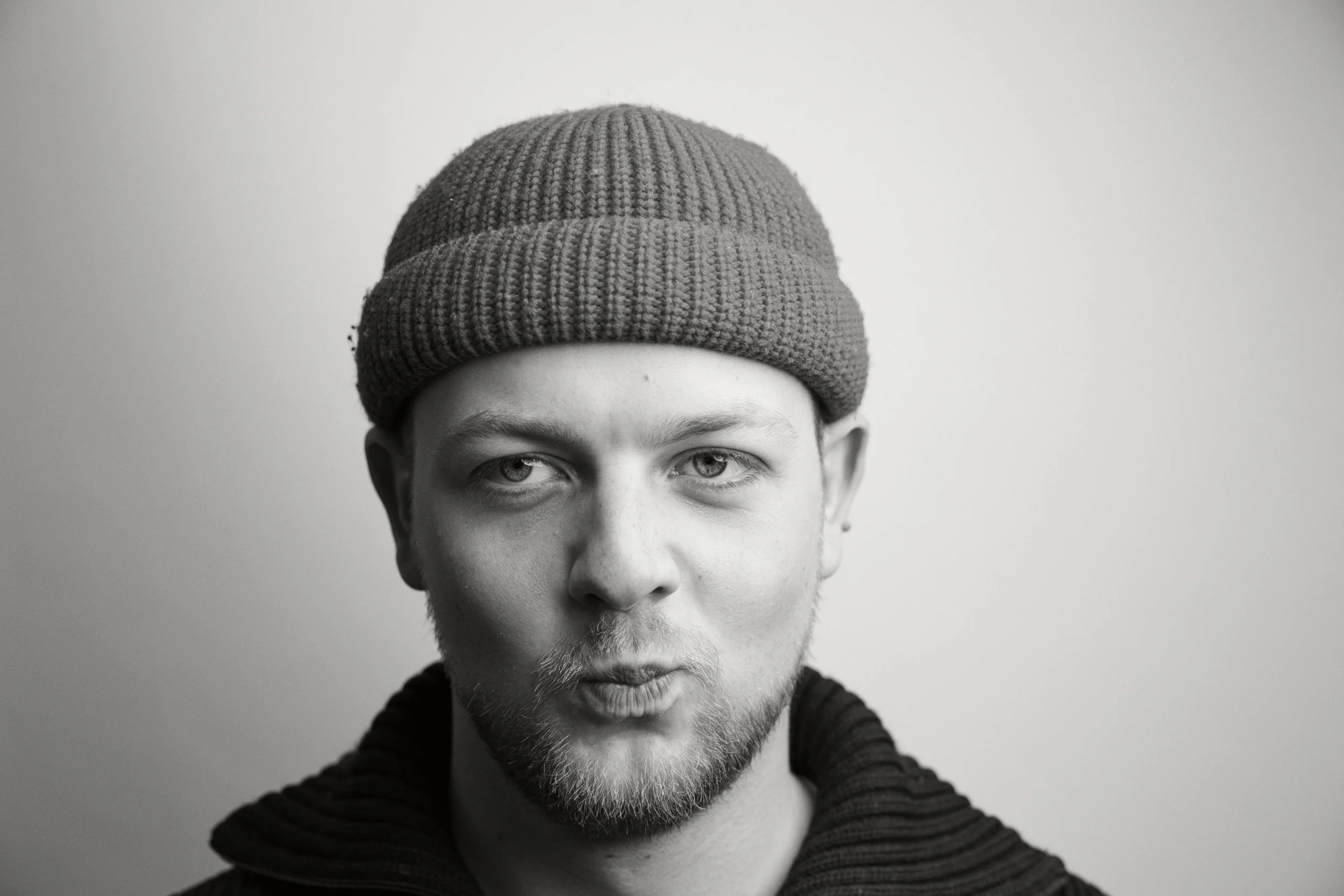 Schwarz-Weiß-Fotografie eines Mannes mit Bart und Tränen auf dem Gesicht, trägt einen grauen Strickmützen und einen schwarzen Pullover, vor hellem Hintergrund.