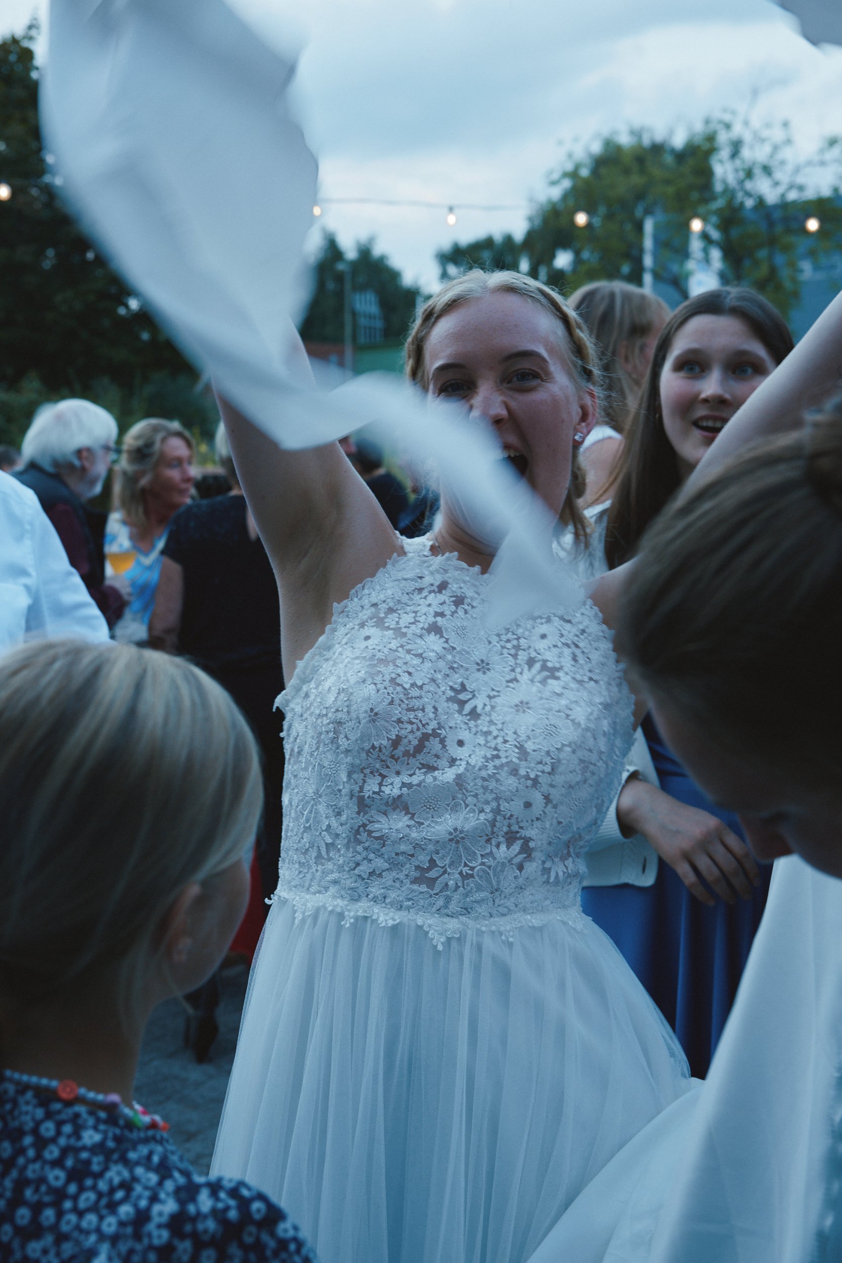 Eine lachende Frau in einem weißen Brautkleid feiert auf einer Outdoor-Party, umgeben von anderen Gästen.