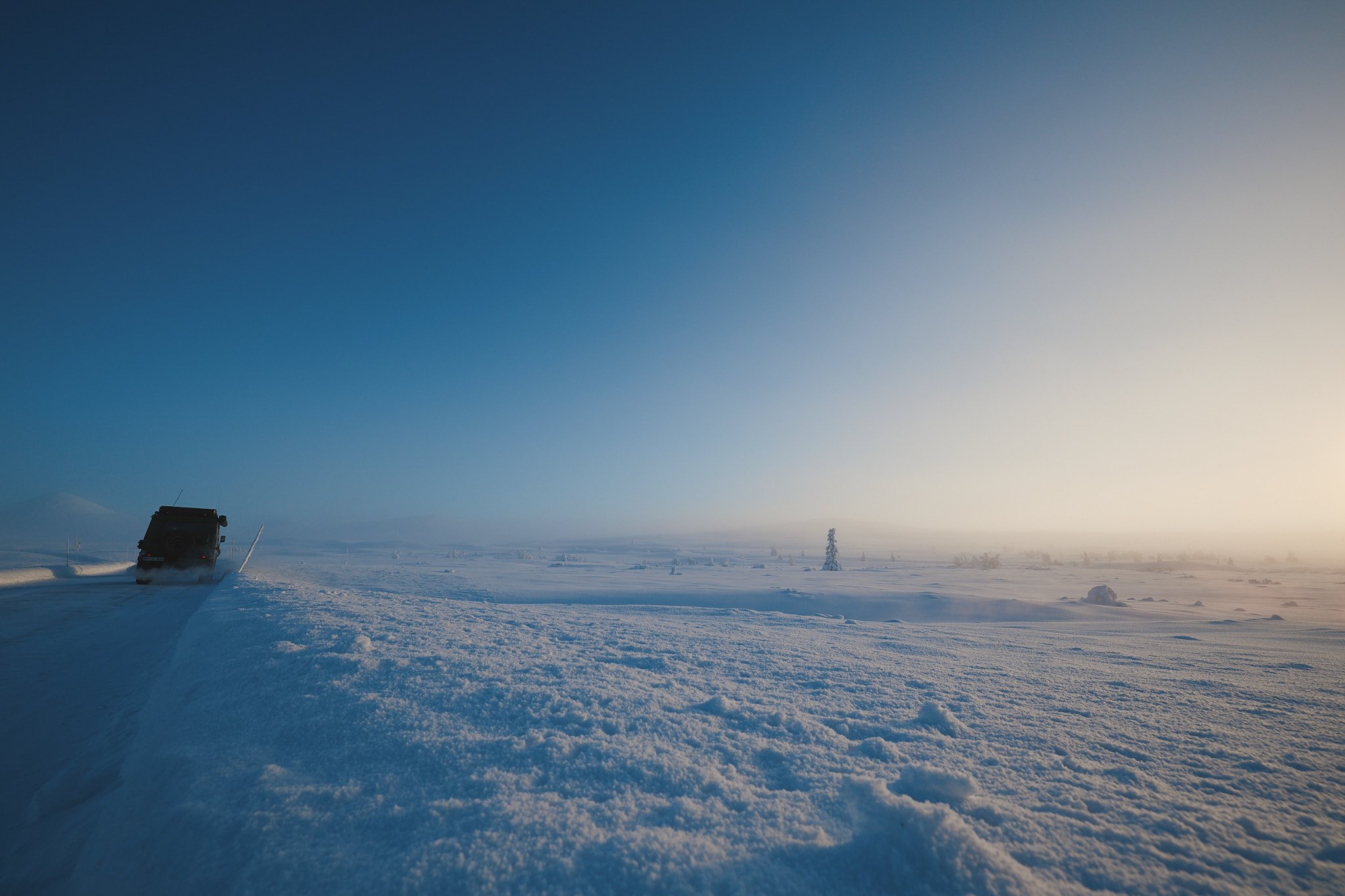 Ein Fahrzeug fährt auf verschneiter Straße durch eine weite Winterlandschaft, unter einem klaren blauen Himmel, mit Bäumen und einer entfernten Bergkulisse im Hintergrund.