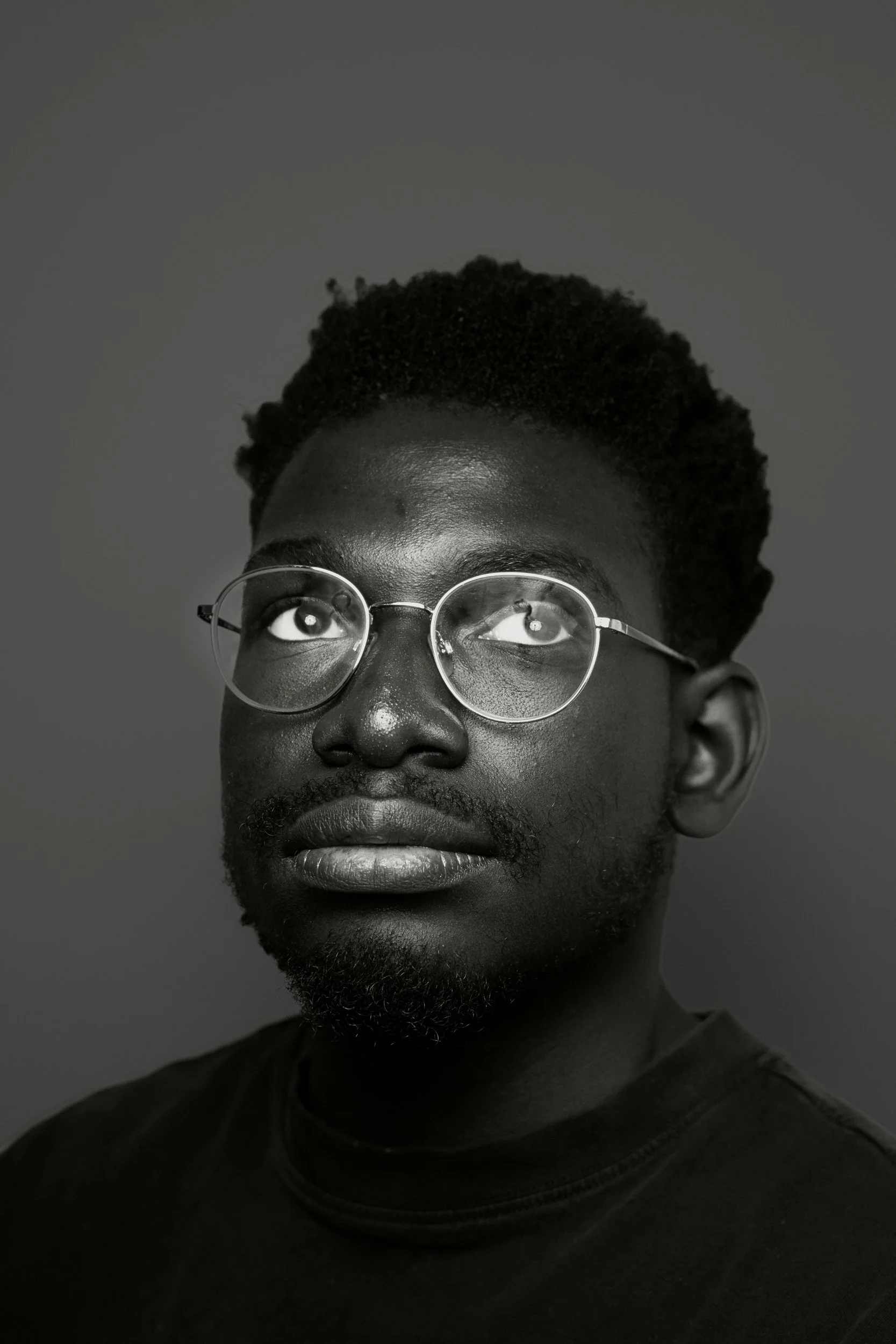 Schwarz-weiß-Fotografie eines Mannes mit Afro-Haartyp, runden Brillen und lockigem Bart, der nach vorne schaut.