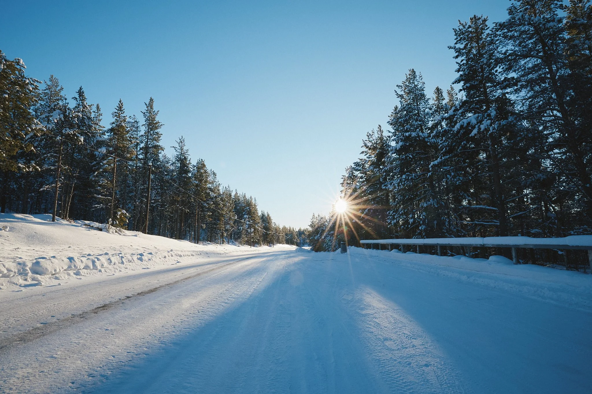 Schneebedeckte Straße im Wald mit Sonnenaufgang oder -untergang am Horizont, klare blauer Himmel.