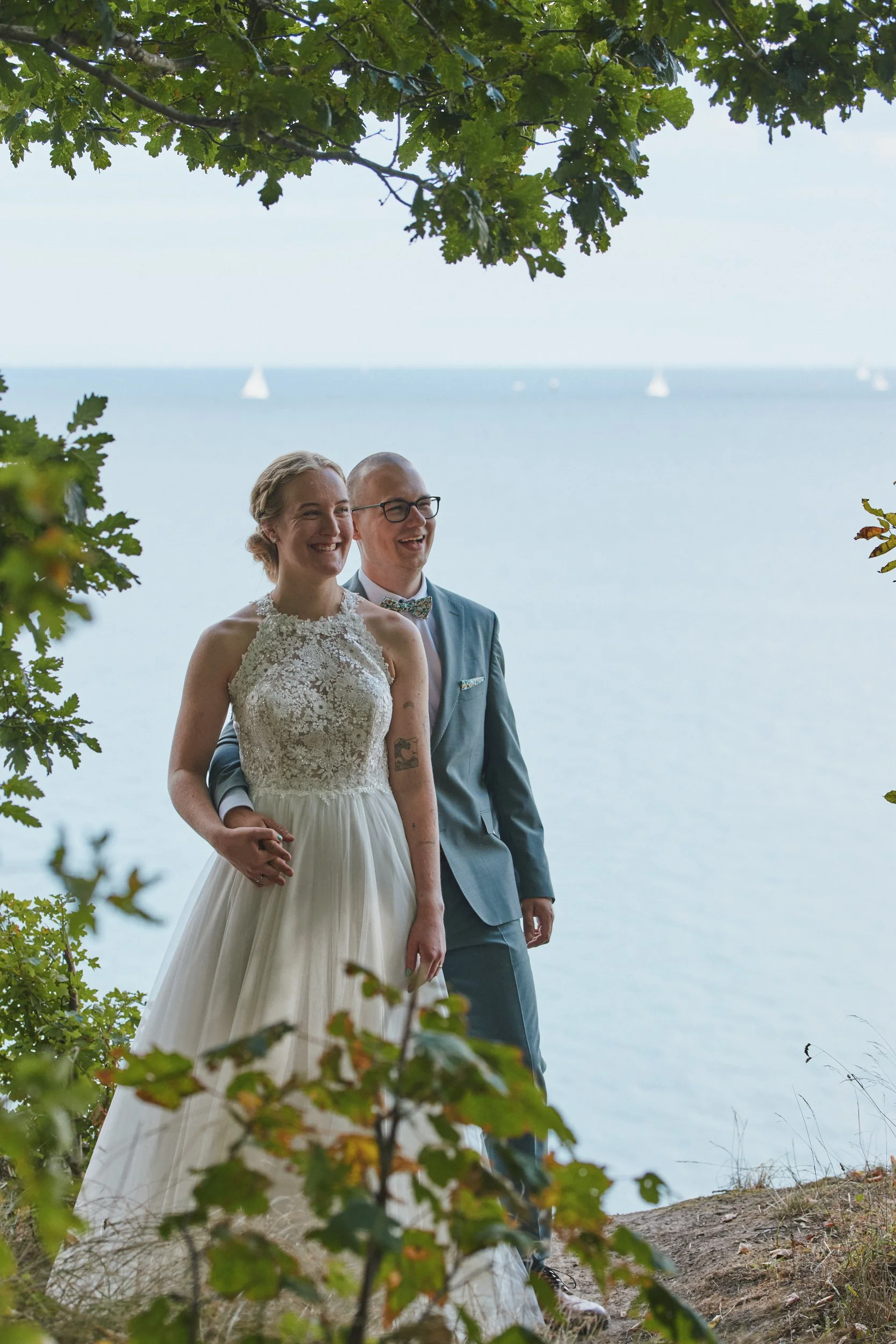 Ein glückliches Paar bei einer Hochzeit am Strand, umgeben von grünen Blättern, mit dem Meer im Hintergrund und Segelbooten am Himmel.