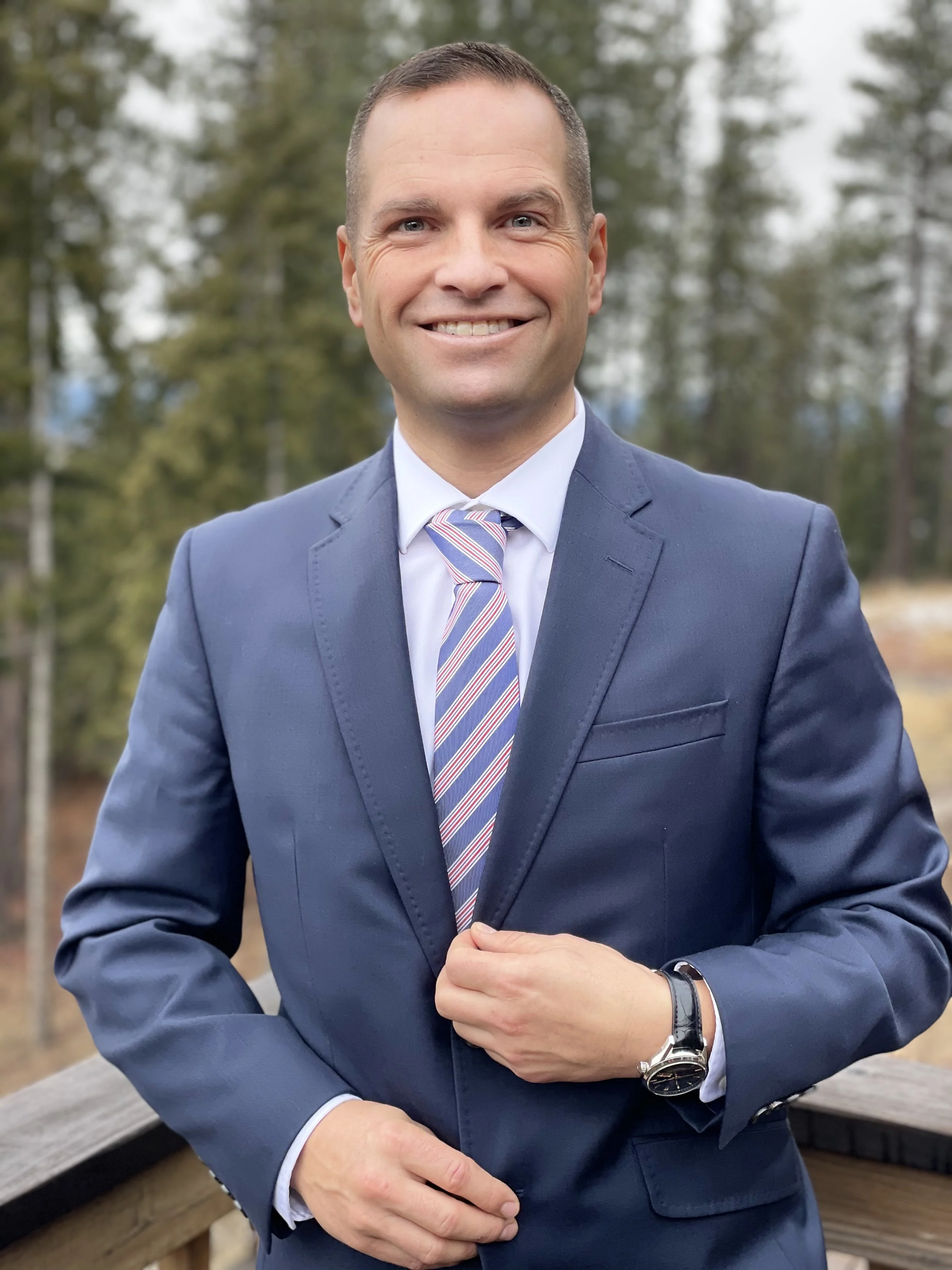 A smiling man in a blue suit, white shirt, and striped tie standing outdoors on a wooden balcony with trees in the background.