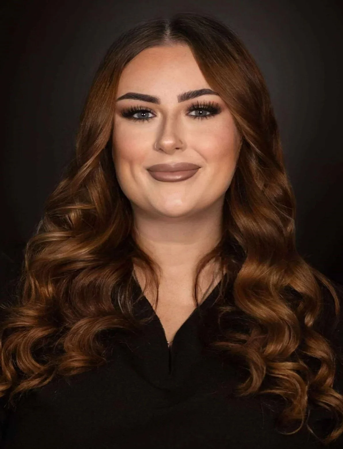Headshot of a woman with long, wavy, auburn hair, smiling slightly, wearing makeup, and dressed in a black top against a dark background.