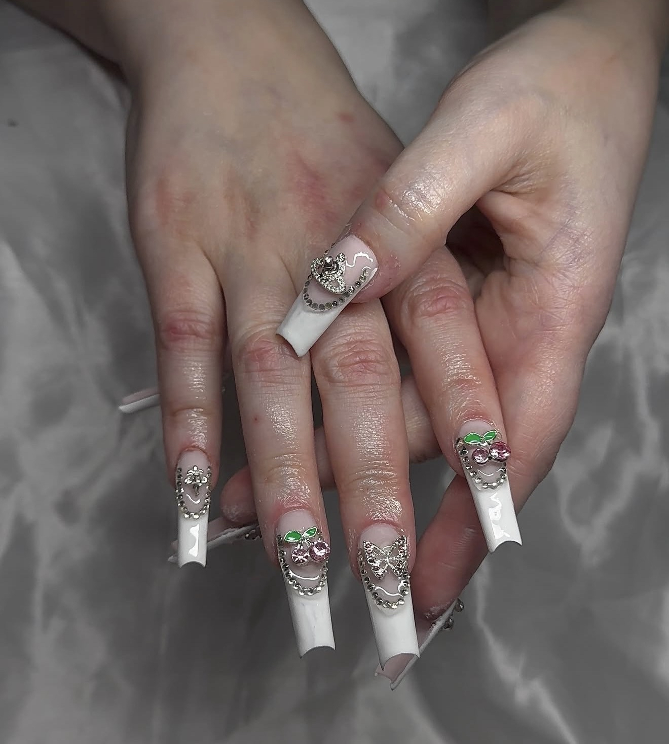 Close-up of a person's hands with elaborate white nail art decorated with rhinestones and 3D embellishments, including butterflies and cherries, against a blurred background.