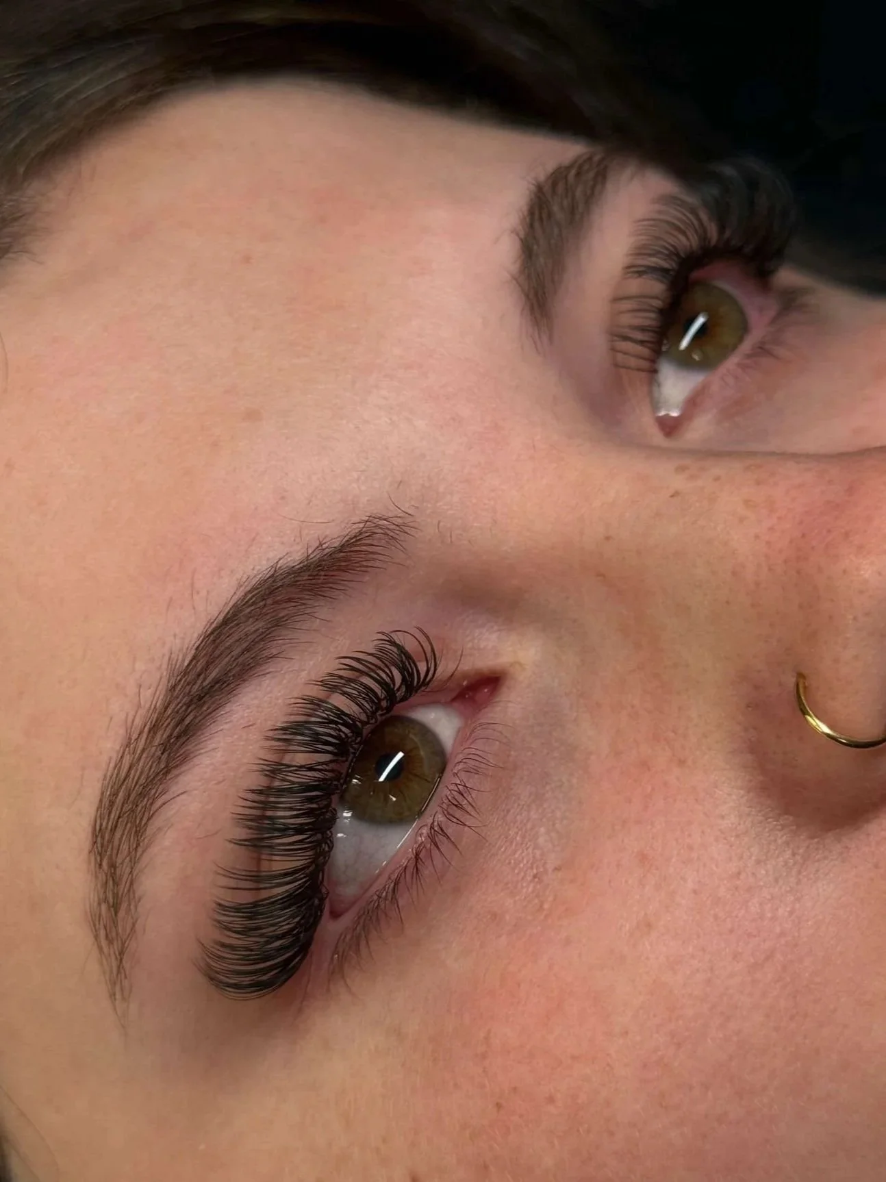 Close-up of two faces lying side by side with their eyes open, showing brown eyes, well-groomed eyebrows, long eyelashes, and gold nose rings.
