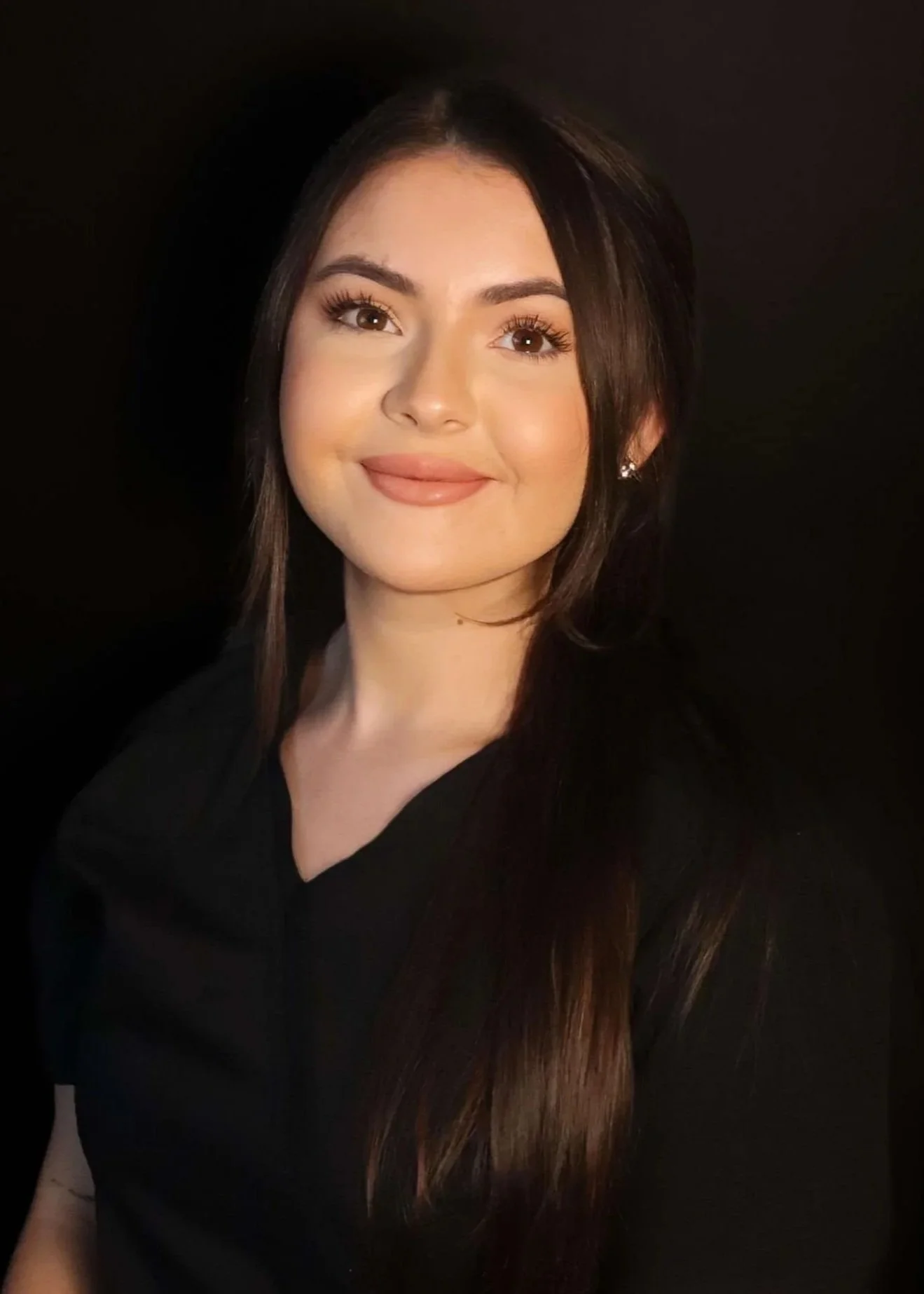 A young woman with long dark brown hair, wearing a black top, smiling against a black background.