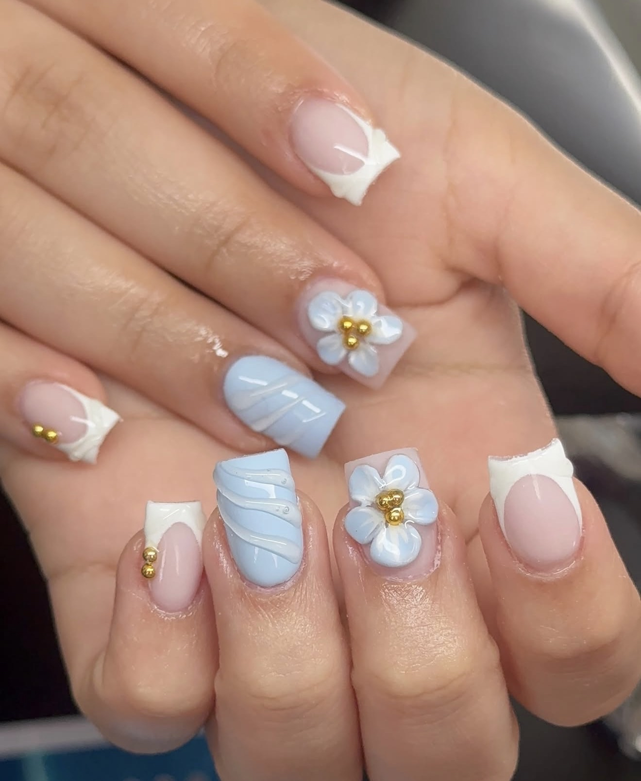 Close-up of hands with manicured nails featuring blue and white designs, gold beads, and 3D white flower embellishments.