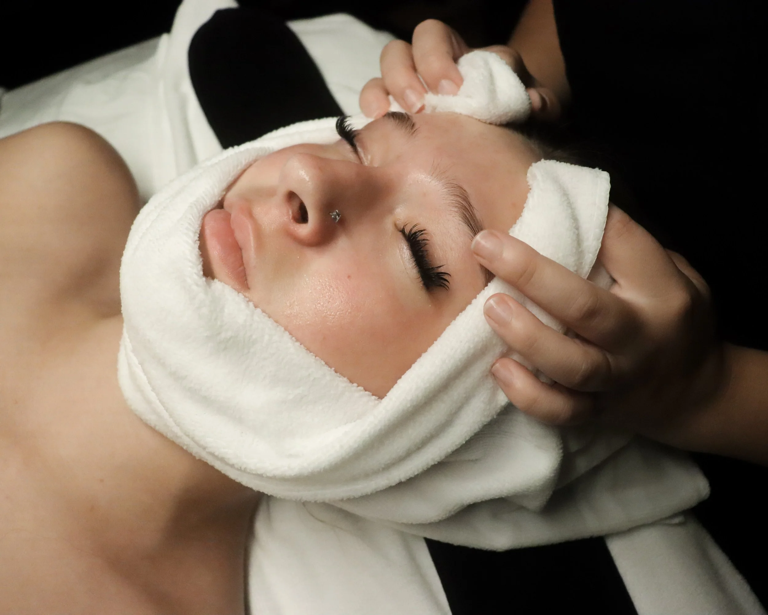 A woman with earrings getting a facial treatment, with a towel wrapped around her head, lying with her eyes closed and a towel on her forehead.