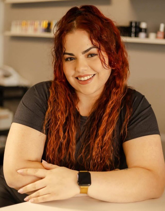 A woman with long, curly red hair smiling and sitting with arms crossed on a table in an indoor setting.