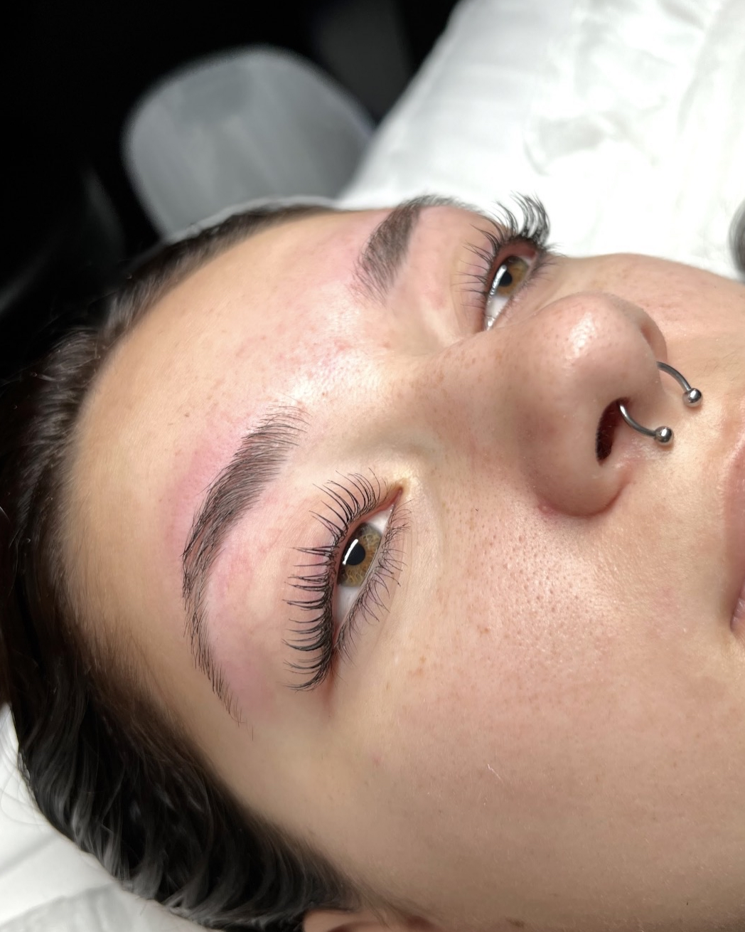 Close-up of a woman lying down, showing her face with freshly done eyelash extensions, thick eyebrows, and a nose piercing with a horseshoe-shaped ring.