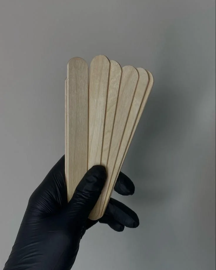 A hand wearing a black glove holding a set of five beige wooden popsicle sticks against a plain gray background.