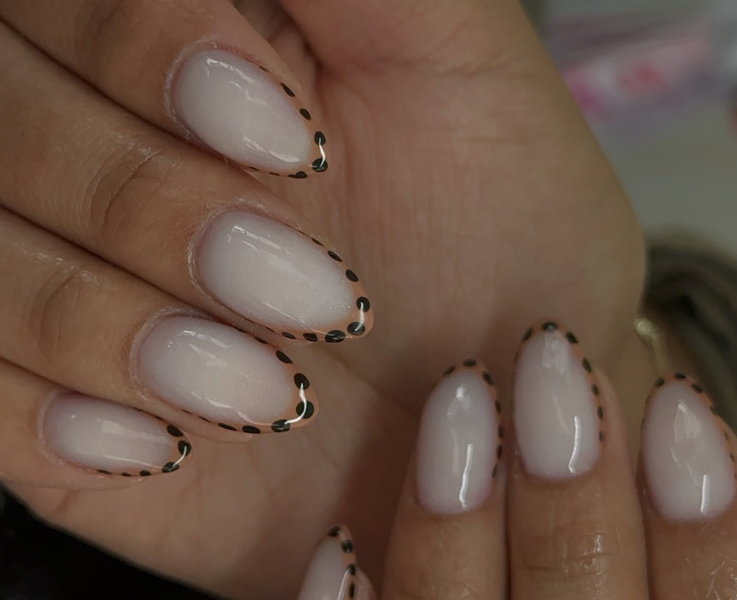 Close-up of a person's hand with neatly manicured nails painted in a light color, featuring black dotted and white accent designs on the French tips.