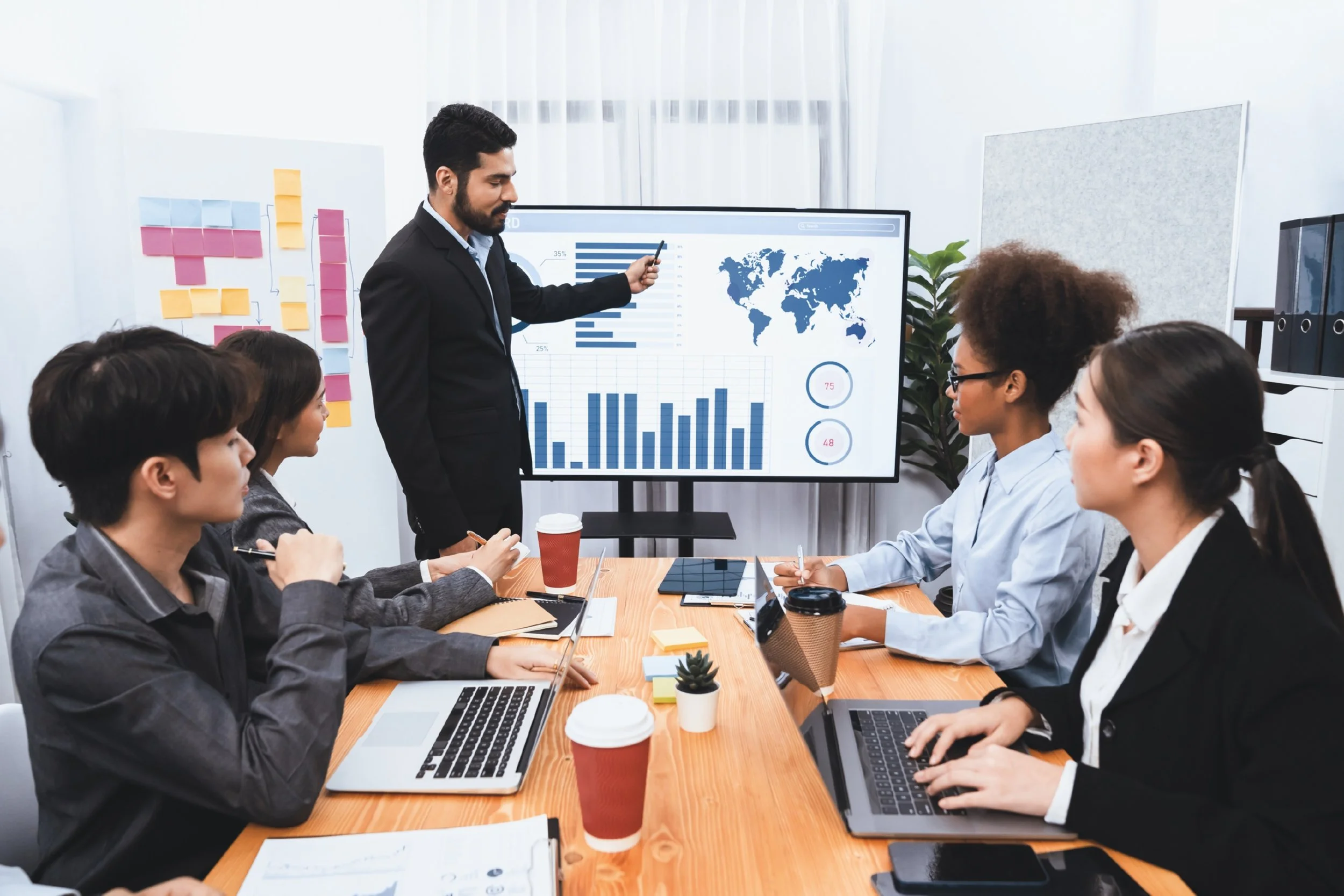 A business meeting with a man presenting data on a large screen with charts, graphs, and world maps to a diverse group of seated professionals at a wooden conference table.