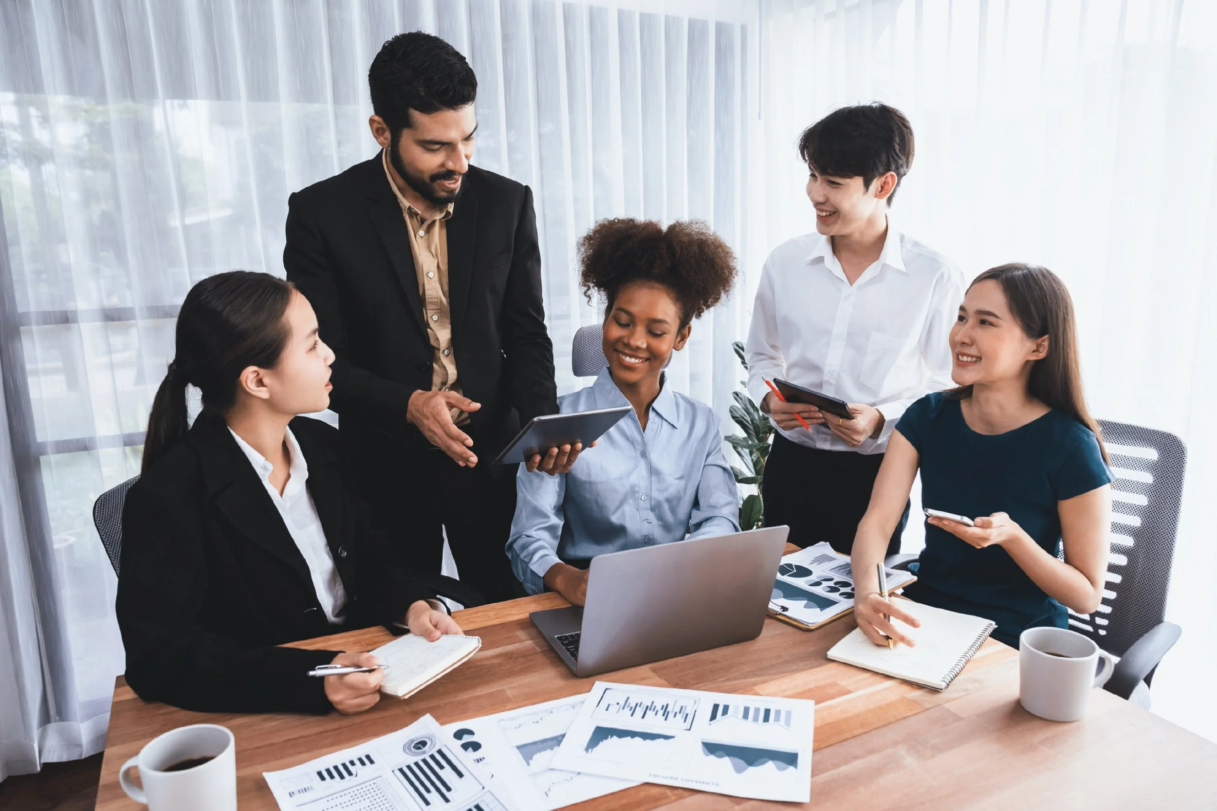 Five diverse professionals in a business meeting in a well-lit office, with documents, a laptop, and coffee cups on the table.