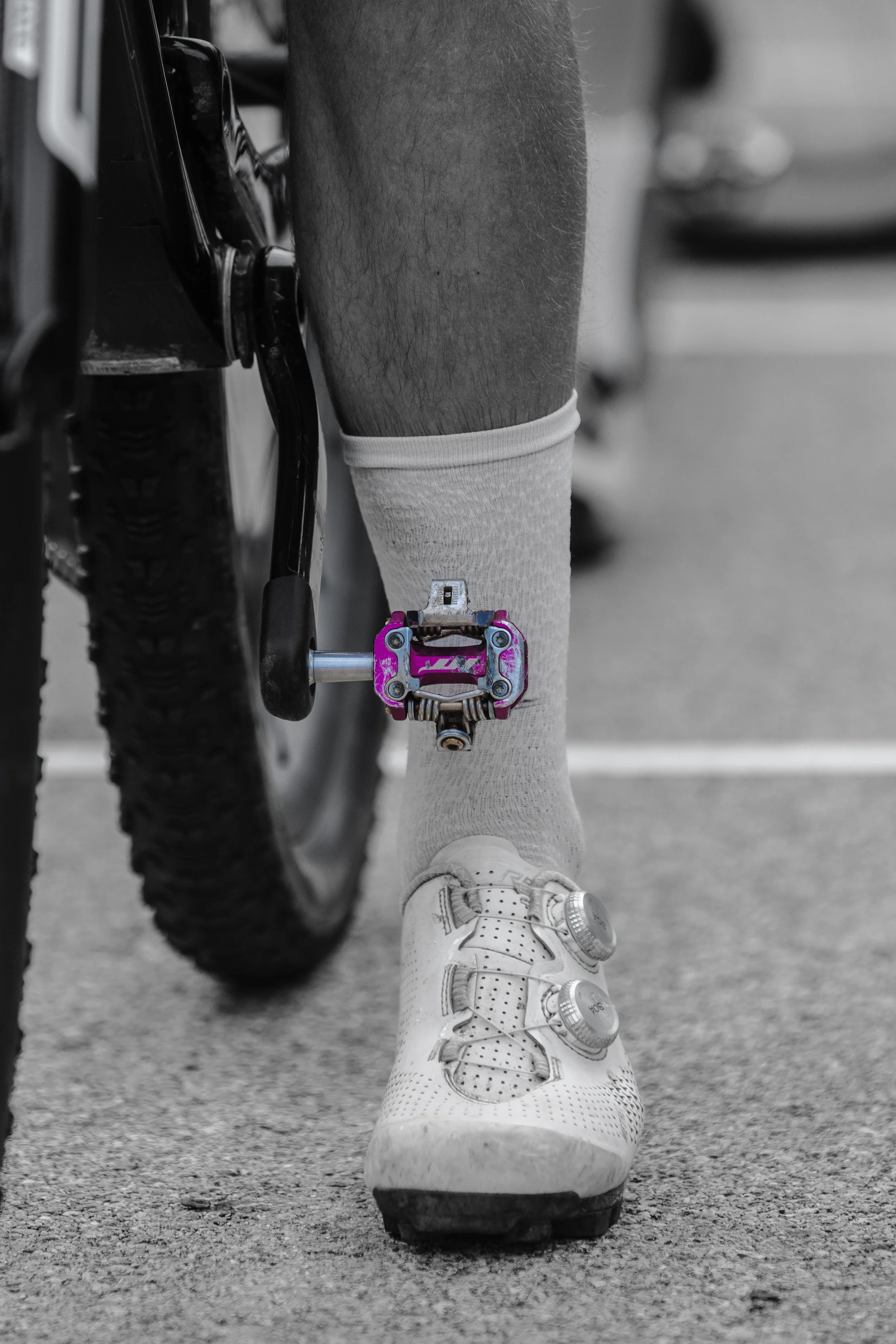 Close-up of a cyclist's foot on a pedal, wearing a white cycling shoe, sock, and part of a bicycle with a black tire visible.