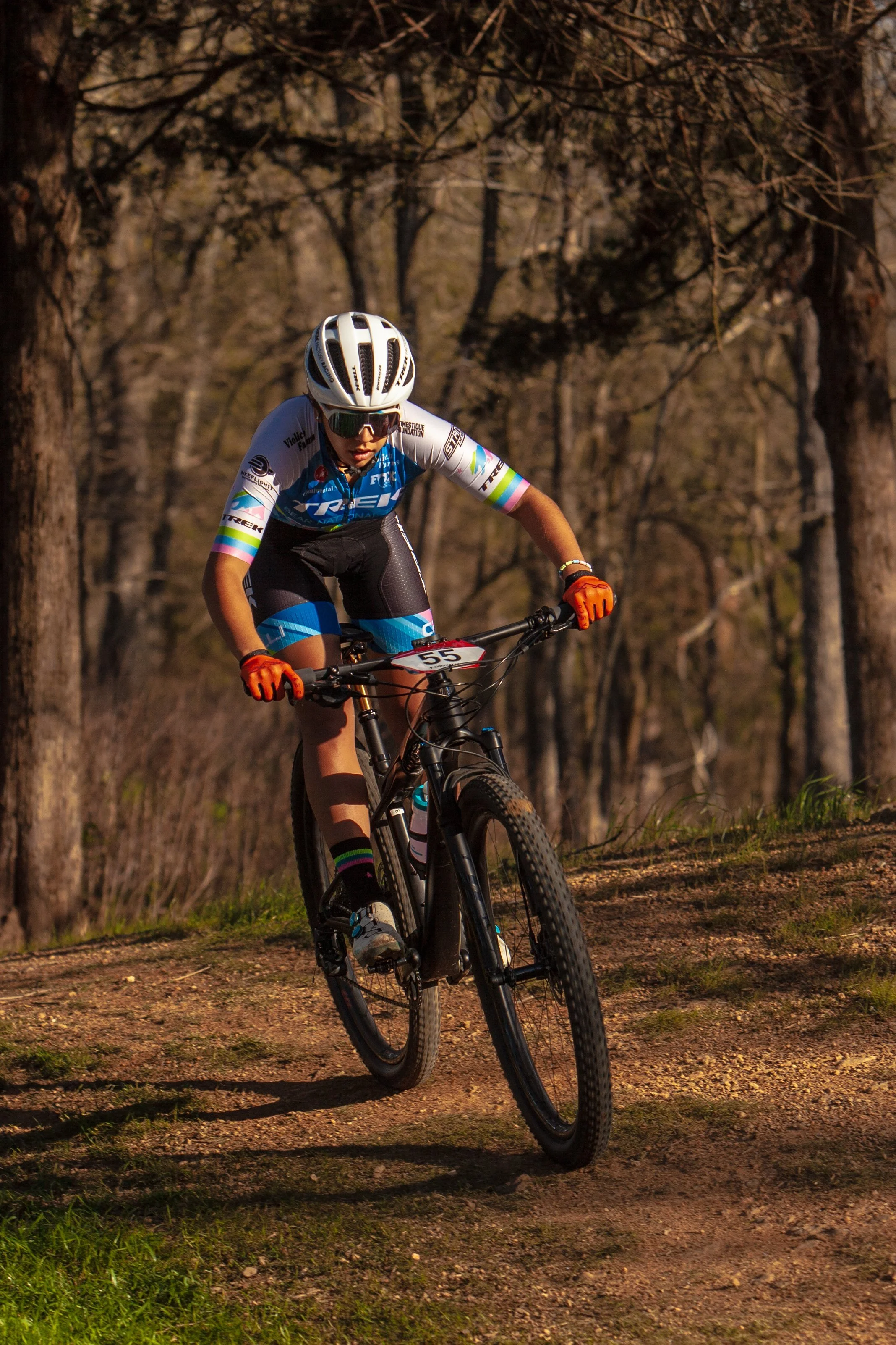 A female cyclist wearing a white helmet, sunglasses, orange gloves, a blue and black cycling outfit, and black shoes riding a mountain bike on a dirt trail through a wooded area.