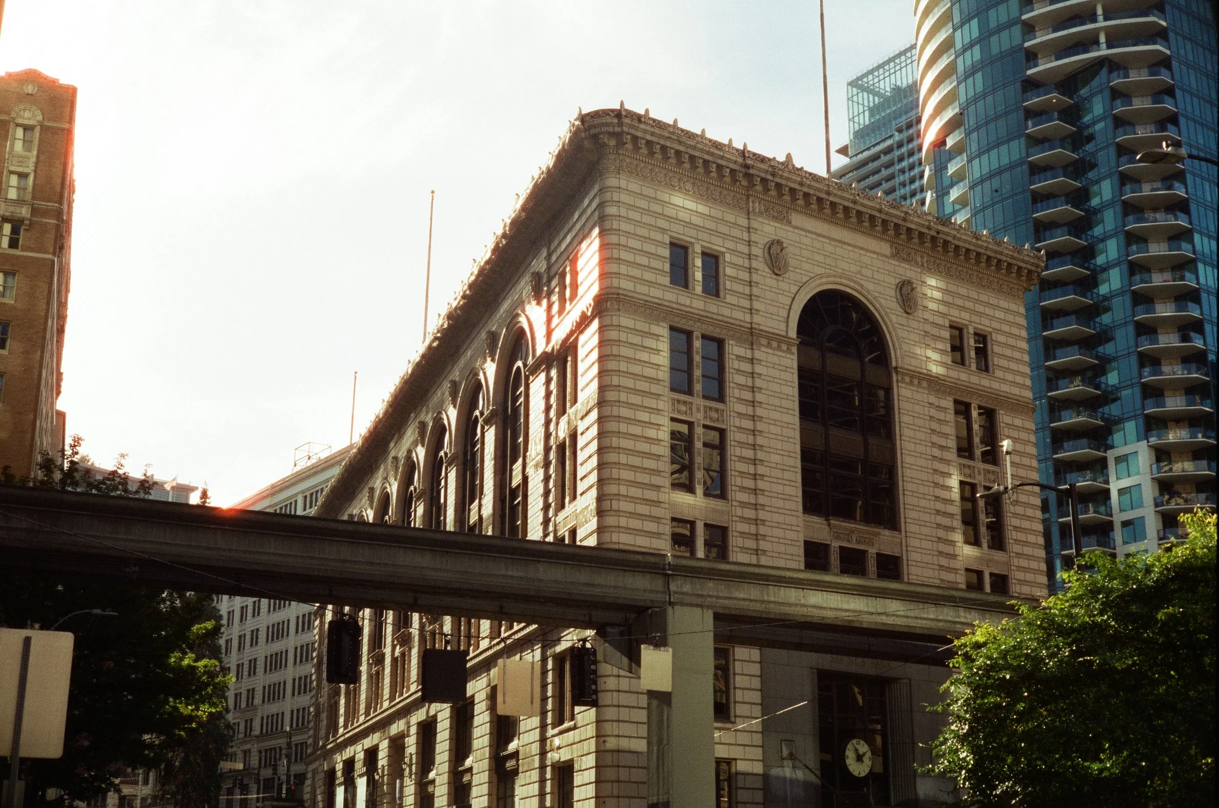 A historic stone building with arched windows and intricate architectural details, with modern high-rise buildings in the background, and a clear sky above.