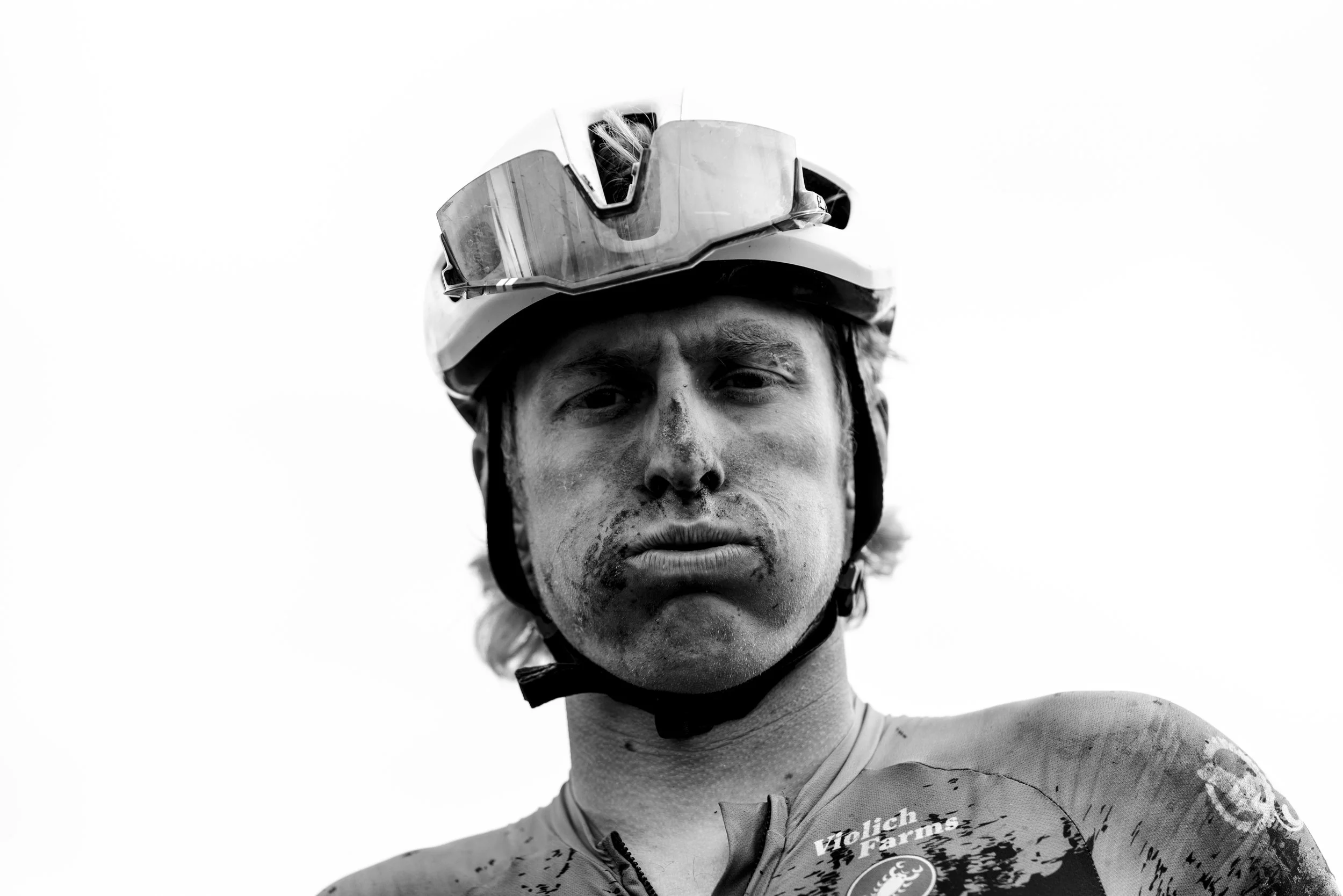 Close-up of a male cyclist with dirt and mud on his face, wearing a helmet, looking serious.