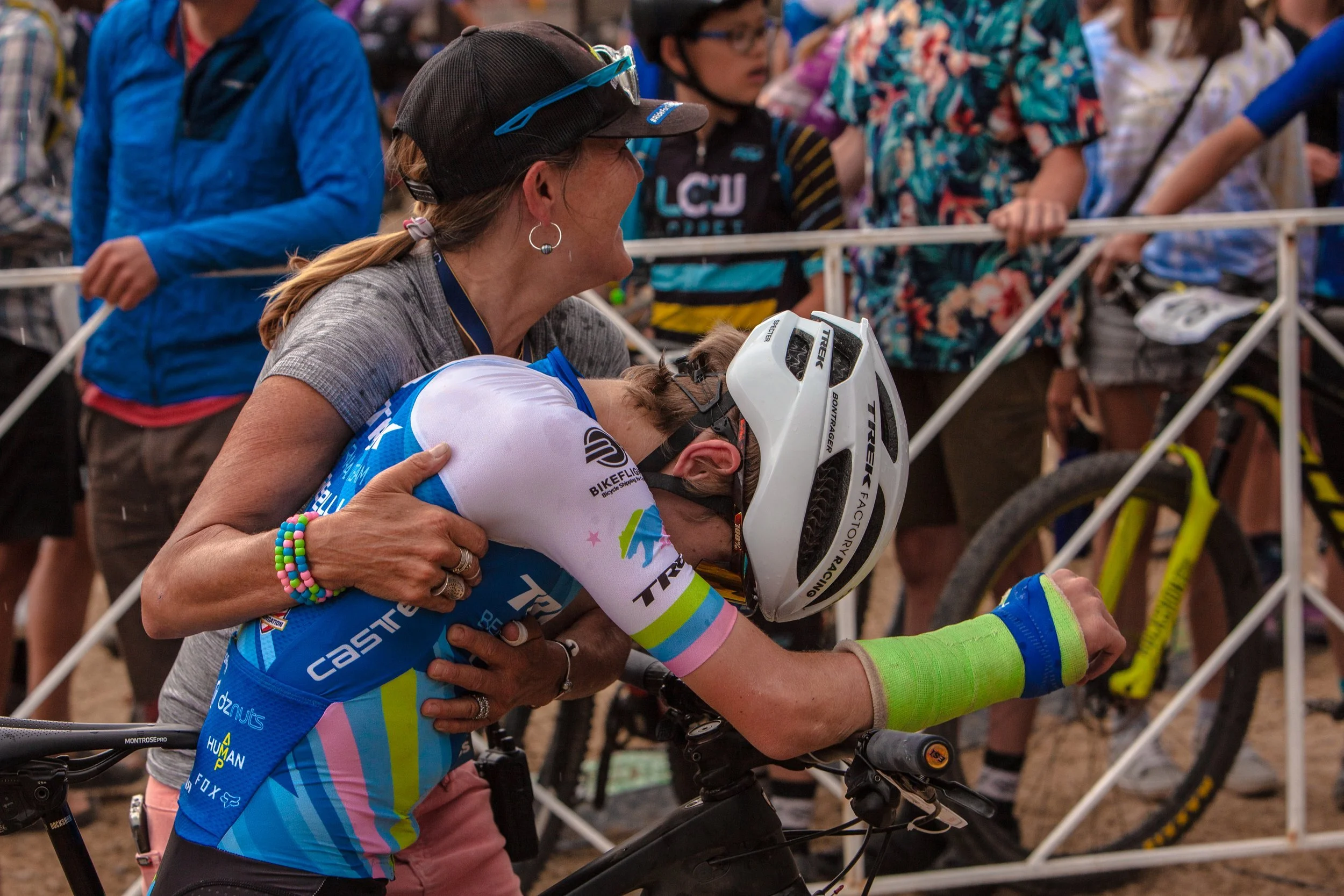 A cyclist with a green sleeve on their right arm is being supported and comforted by a woman during a race or event. The woman is wearing a gray shirt, a black cap, and several colorful bracelets. The cyclist is wearing a white helmet and a blue, pink, and green cycling jersey.