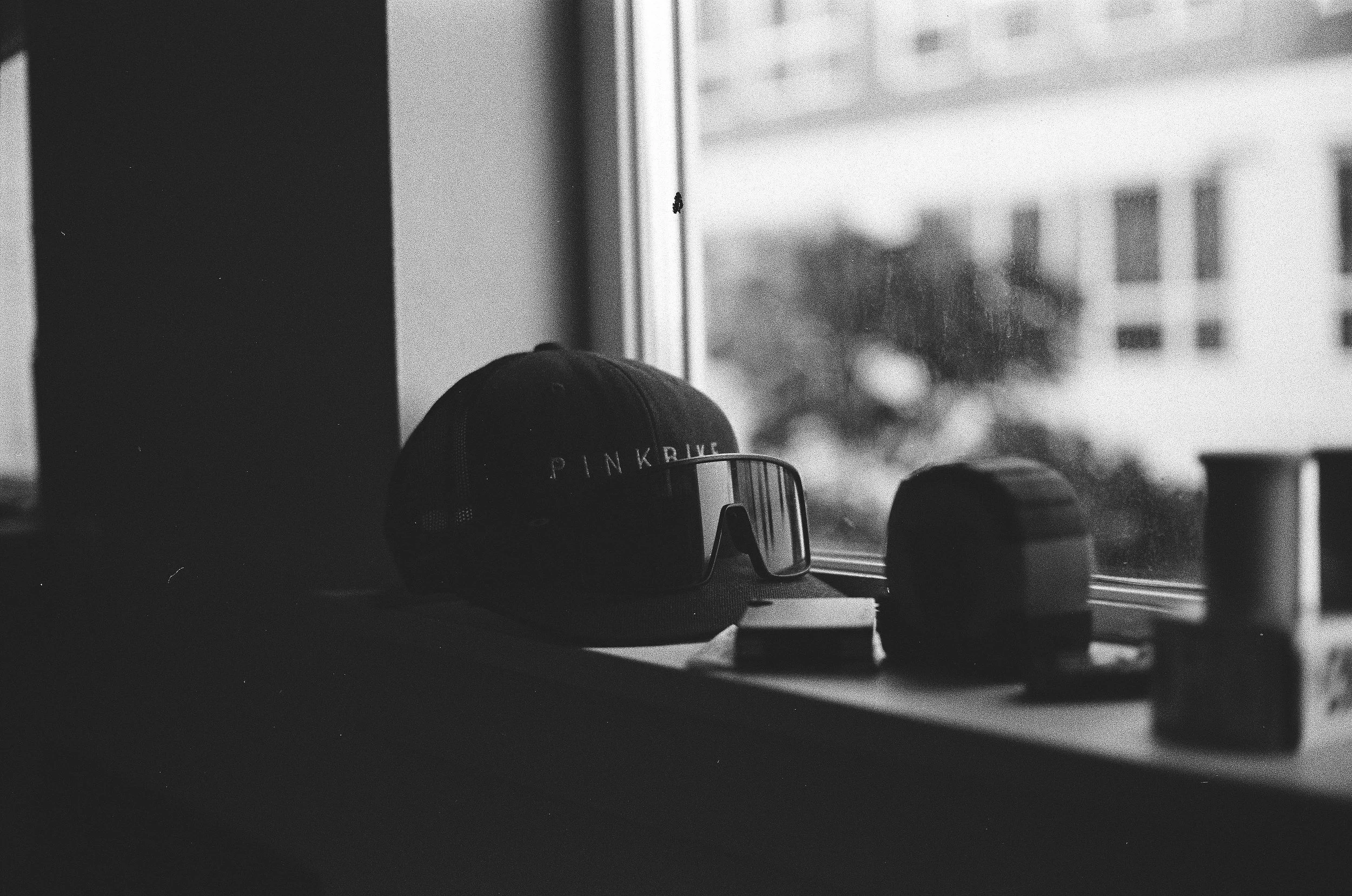 Black and white photograph of a windowsill with a Pinkbike cap, sunglasses, and other small objects, with a blurry cityscape outside the window.