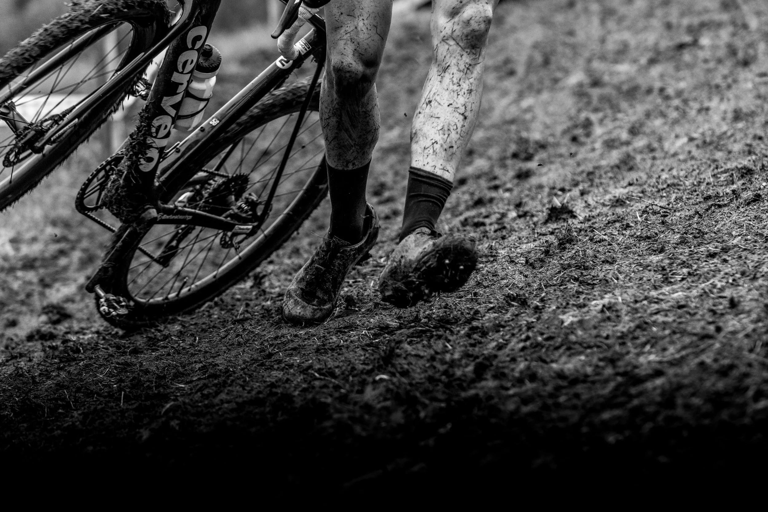 A cyclist with muddy legs and feet falls or slides on a muddy trail next to their cyclocross bike, which is also muddy.