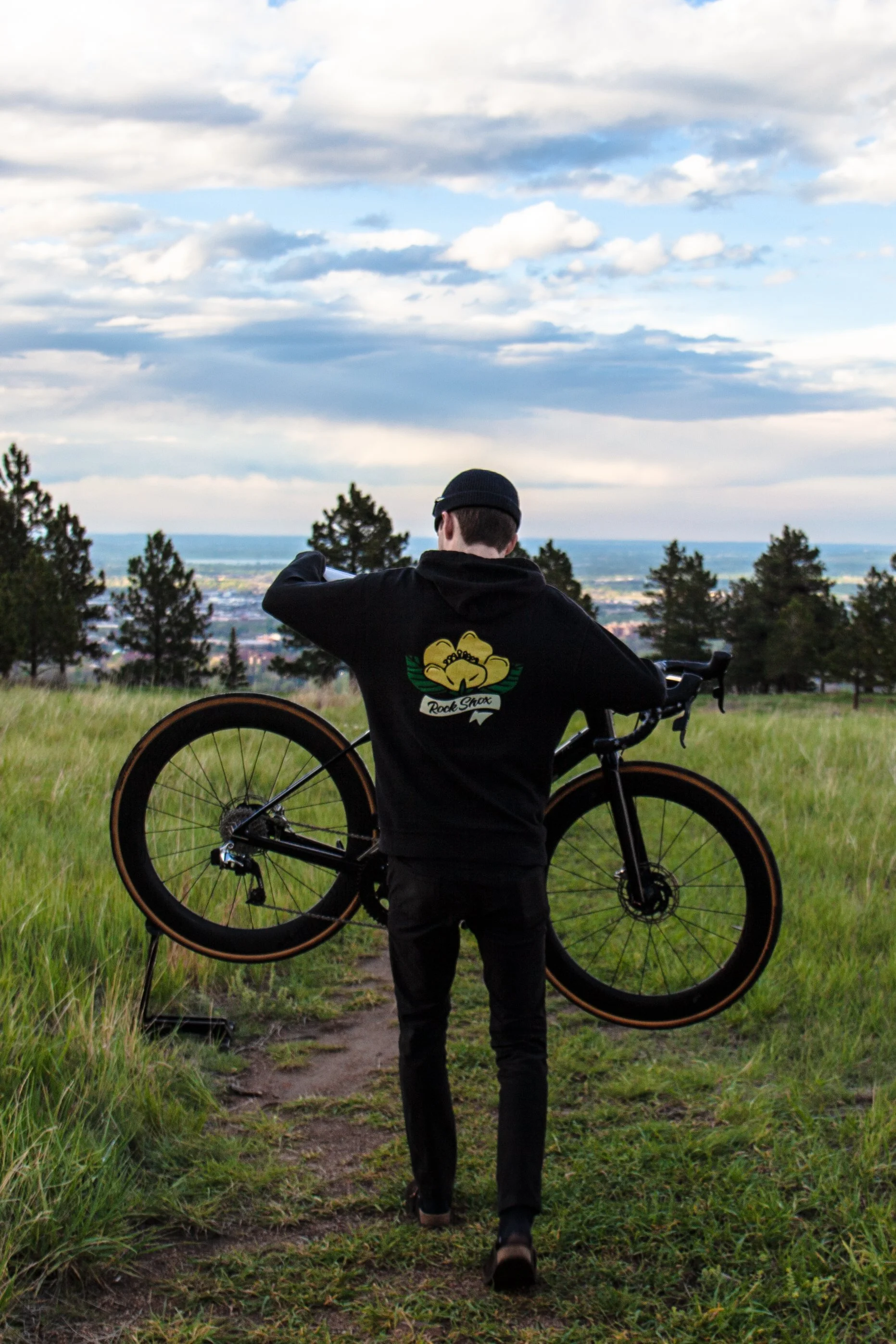 Person walking on grassy trail holding a black bicycle over their shoulder, wearing a black hoodie with a yellow emblem on the back, in a green landscape with trees and a cloudy sky.