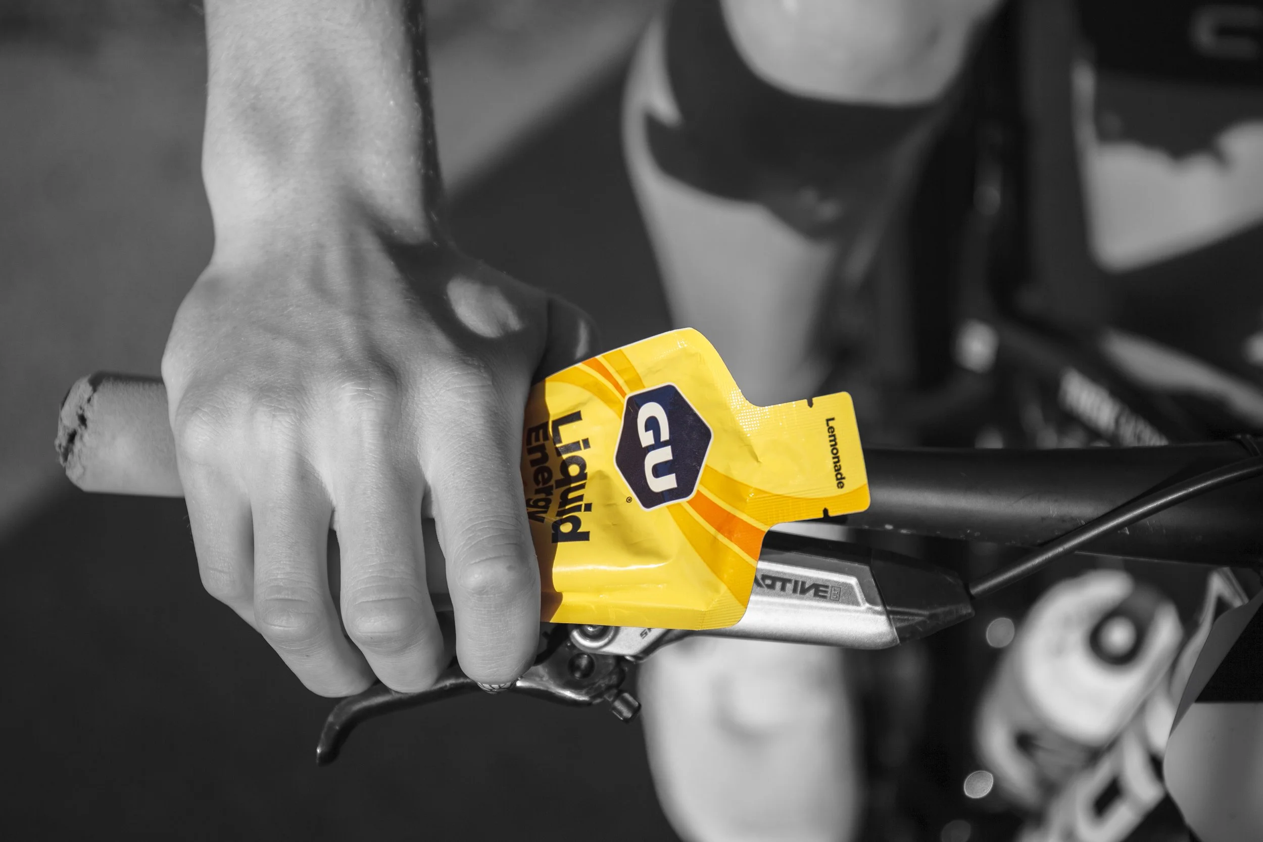 A person's hand gripping a bicycle handlebar with a yellow Gu Liquid Power energy gel package in hand. The image is in black and white except for the yellow energy gel package.