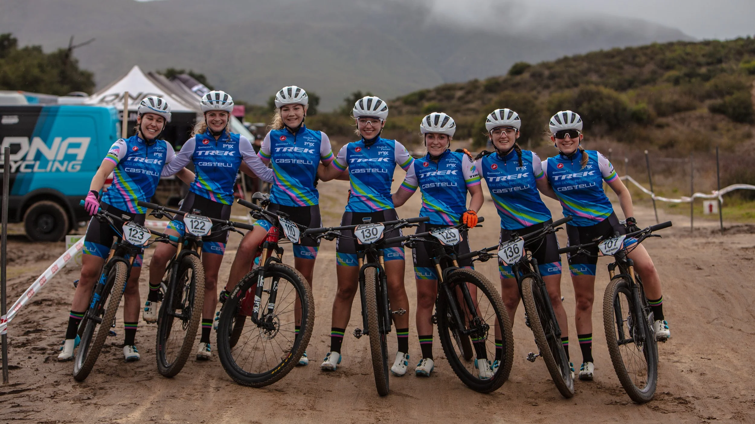 A group of seven female mountain bikers in matching blue jerseys and shorts, wearing helmets and gloves, posing with their bikes on a dirt trail with hills in the background.