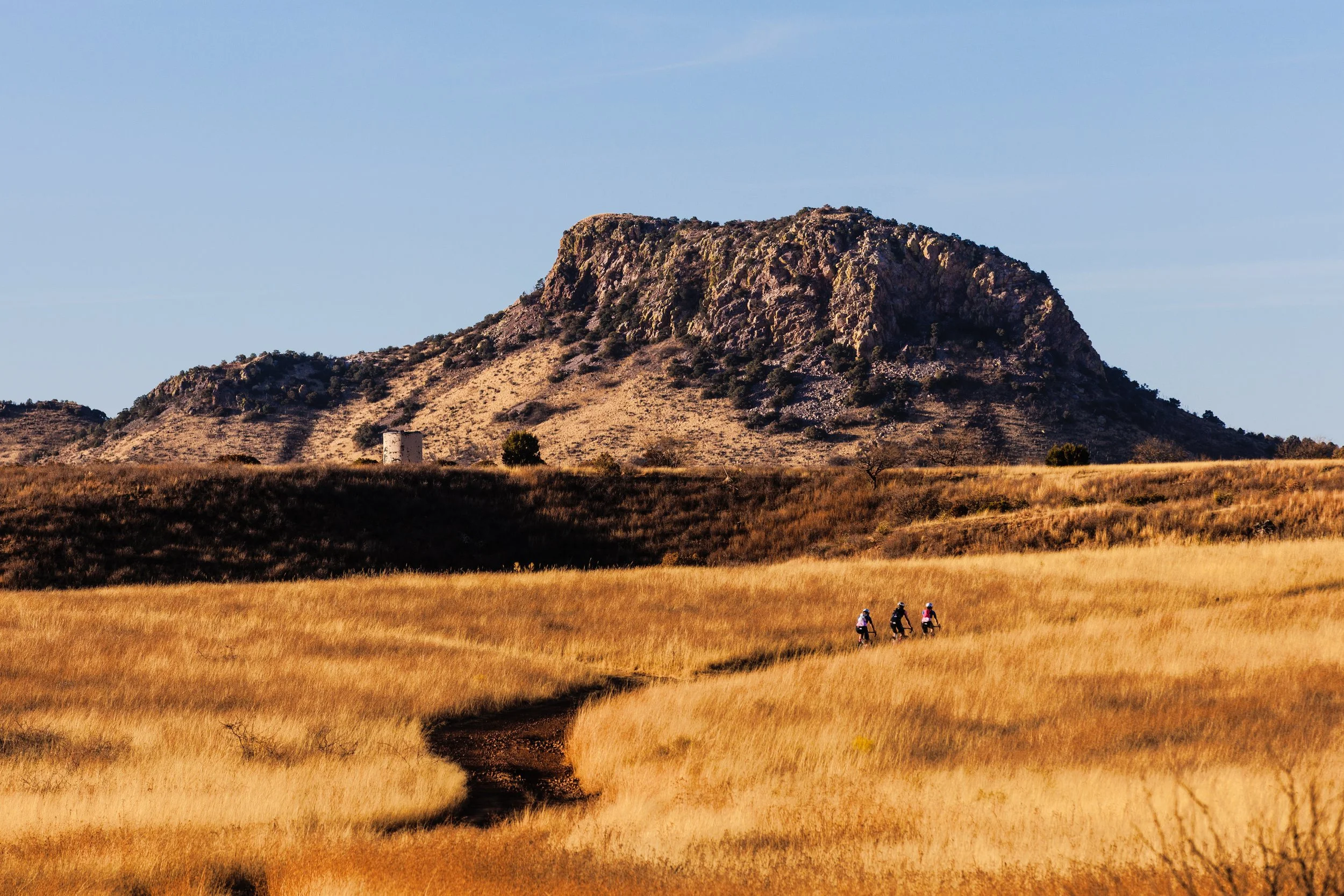 Three people biking on a dirt trail through a golden field with a large rocky hill in the background under a clear blue sky.
