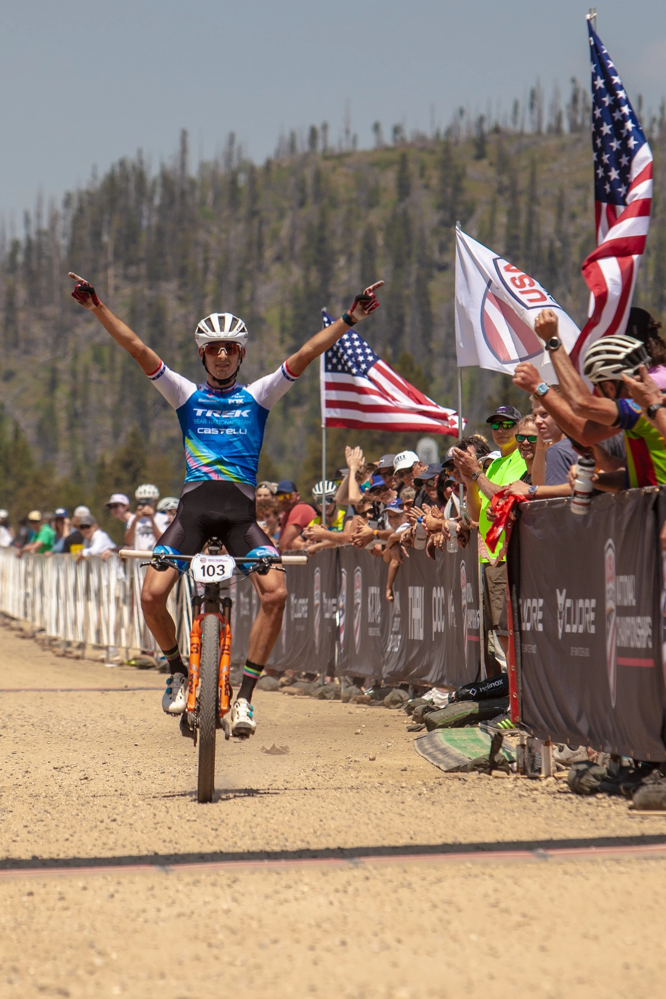 A male cyclist crossing the finish line during a race, lifting his arms in celebration. He is wearing a blue jersey, black shorts, a white helmet, and sunglasses. The crowd on the sides is cheering, holding American flags, and clapping.
