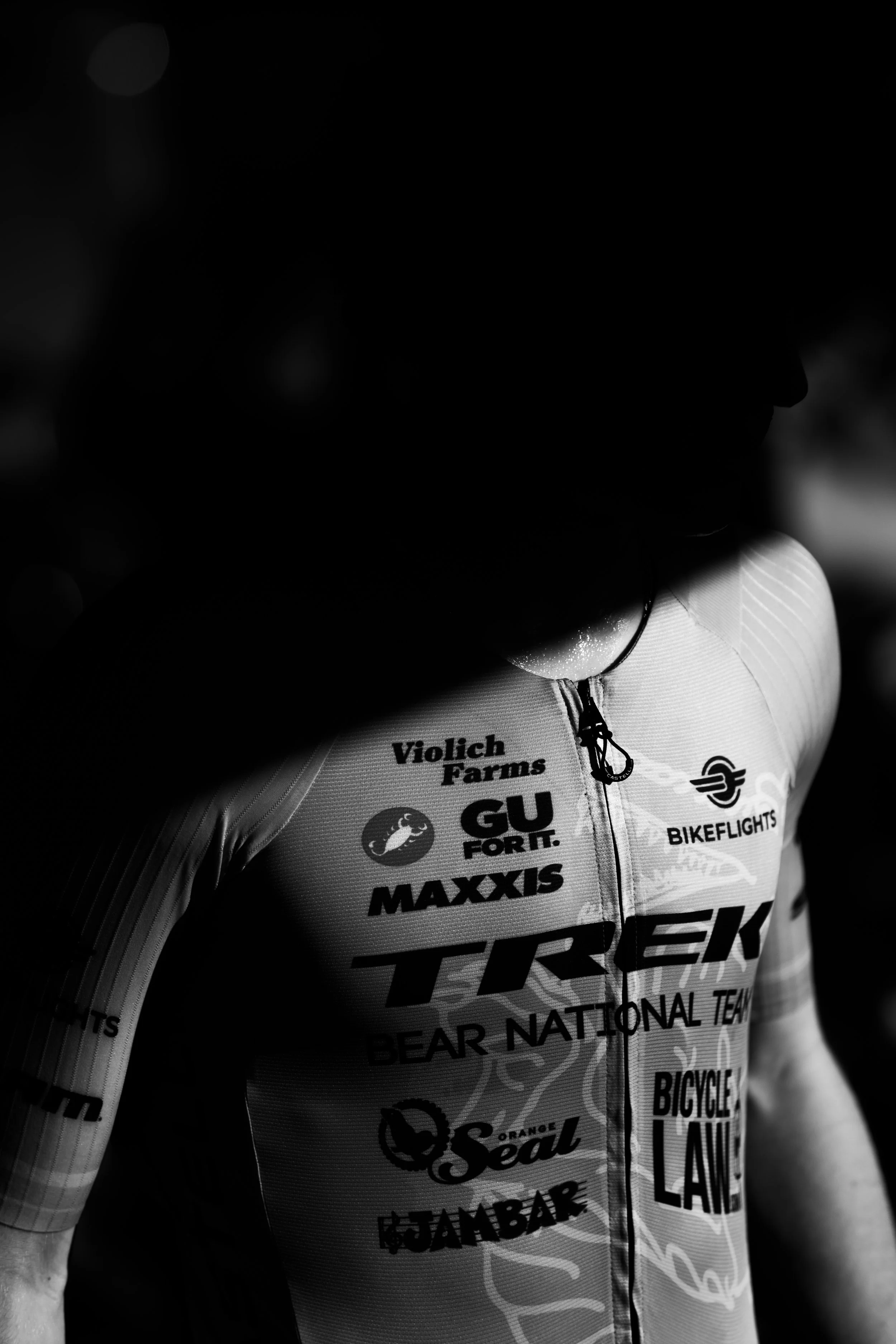 Close-up of a cyclist wearing a jersey with various sponsor logos, with part of their face obscured by shadow.