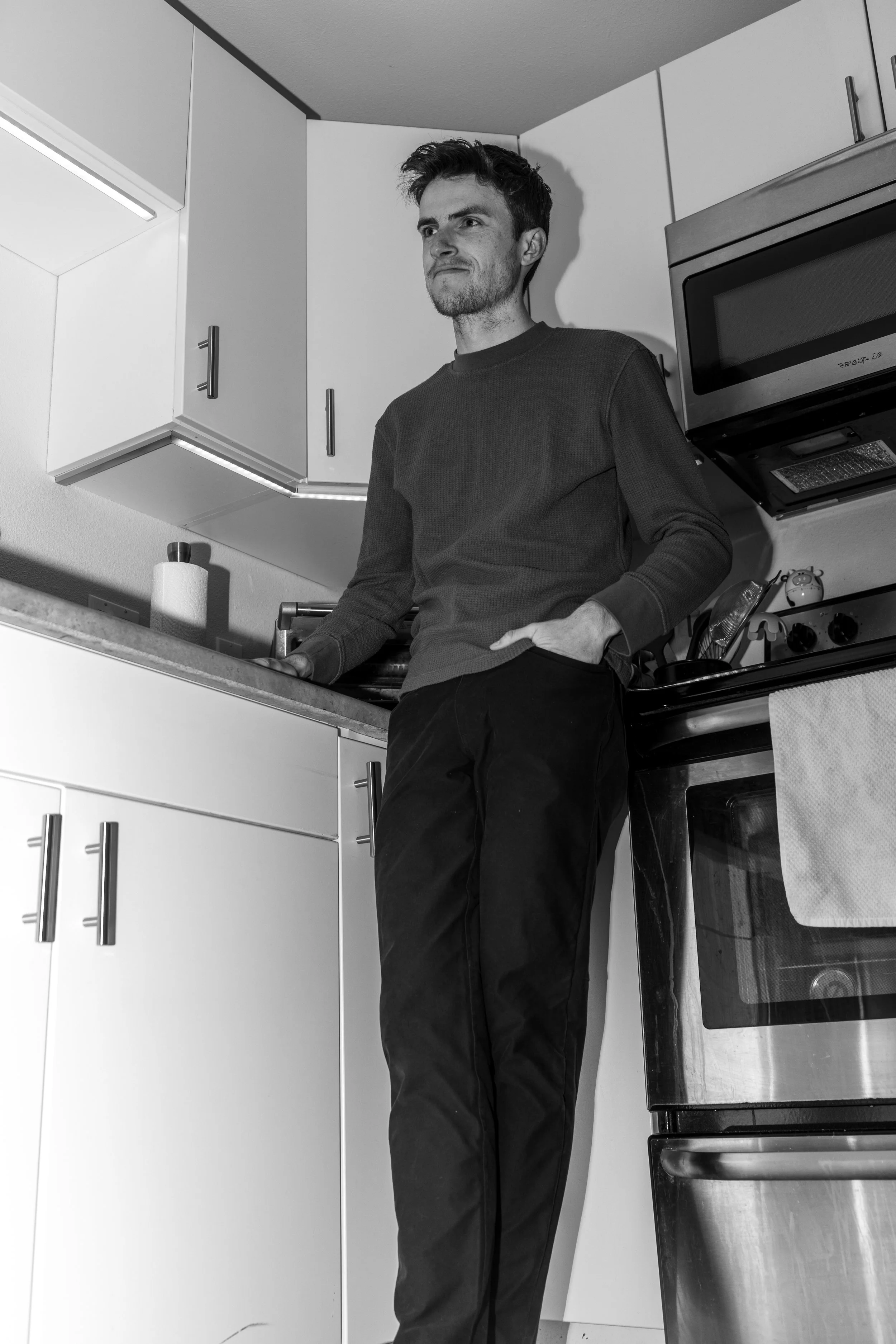 A young man with dark hair and a beard stands in a kitchen, leaning against the countertop with one hand on the counter and the other in his pocket. He is wearing a long-sleeve shirt and dark pants, with a serious expression on his face. The kitchen has white cabinets, a microwave above the stove, and items on the counter including a paper towel roll.