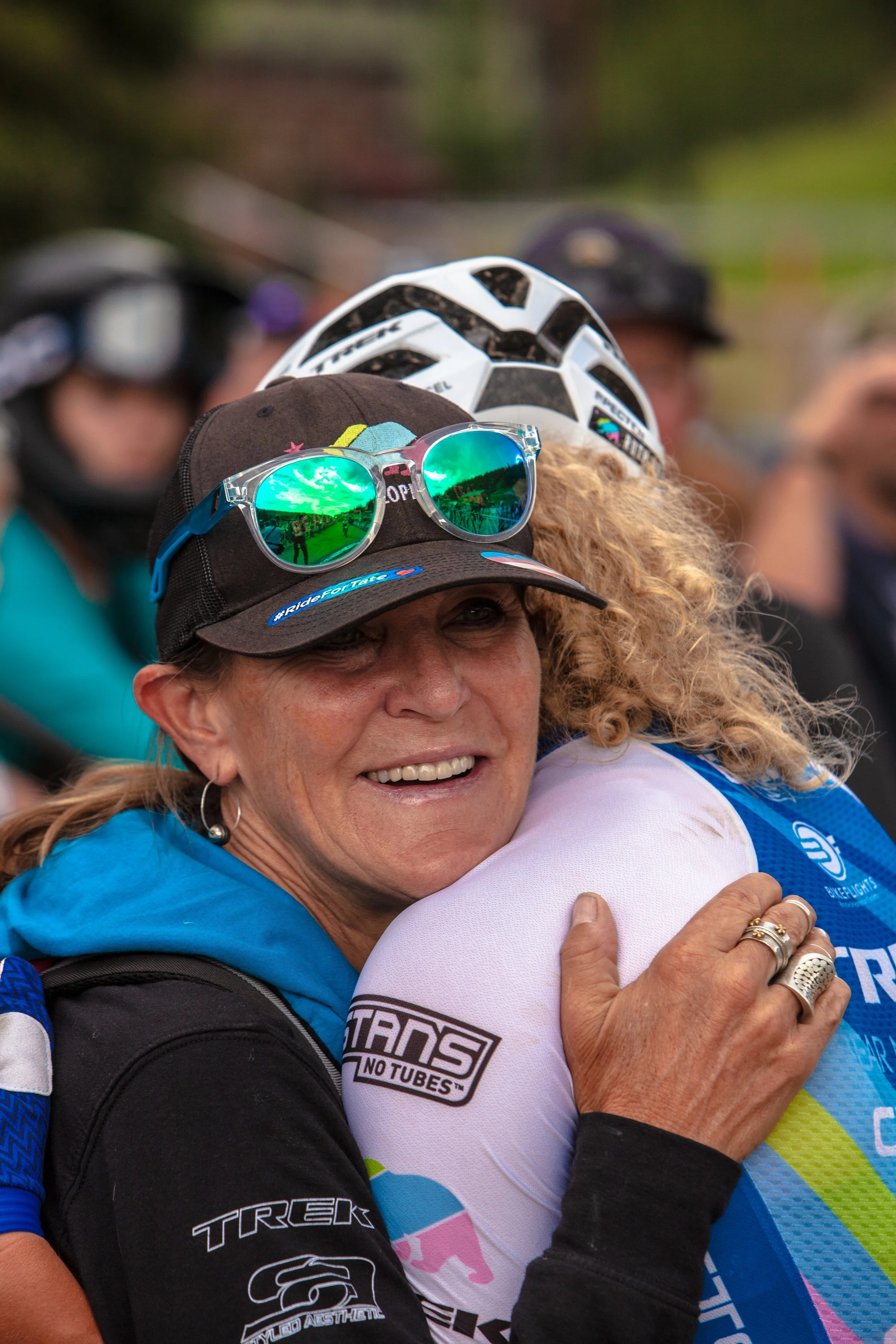 Women hugging, smiling, wearing cycling gear, with helmets and sunglasses, outdoors.