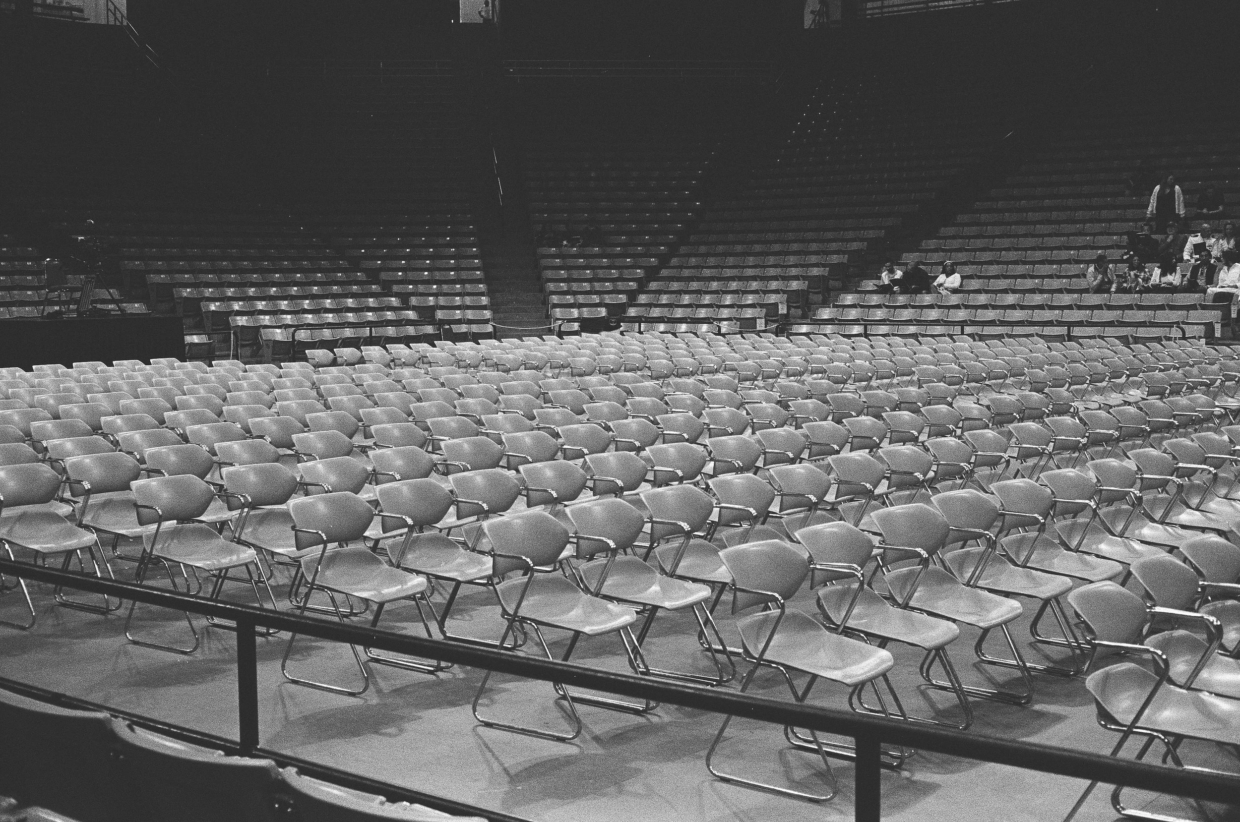 Empty chairs set up in rows inside an auditorium or concert hall, with a few people sitting in the background.
