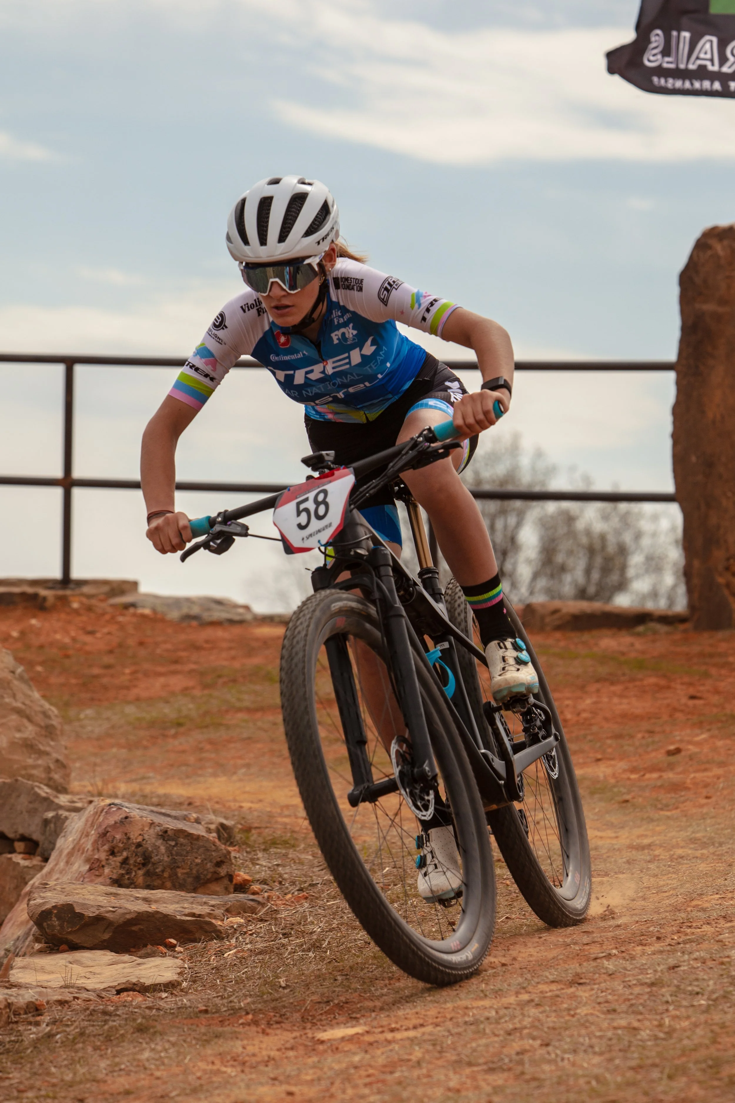 Young female mountain biker wearing a white helmet, sunglasses, and a blue cycling jersey with the number 58 riding downhill on a dirt trail with rocks and a metal fence in the background.