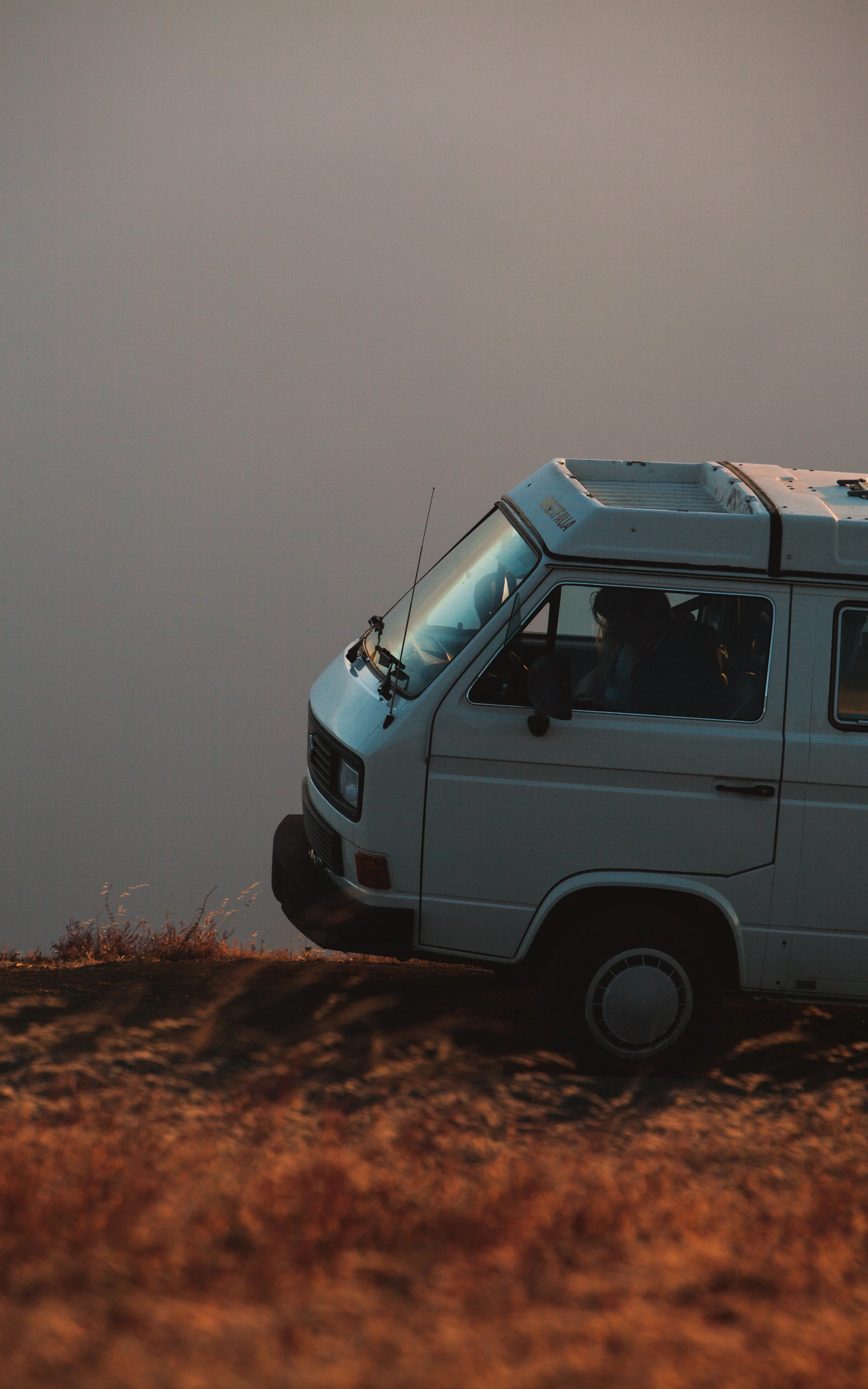 A white camper van parked on a hill during sunset, with the sky behind it and dry grass in the foreground.
