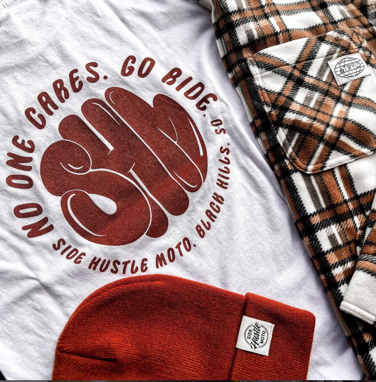 A white T-shirt with a red graphic and text, a pair of plaid-patterned pants, and a red knit hat laid flat.
